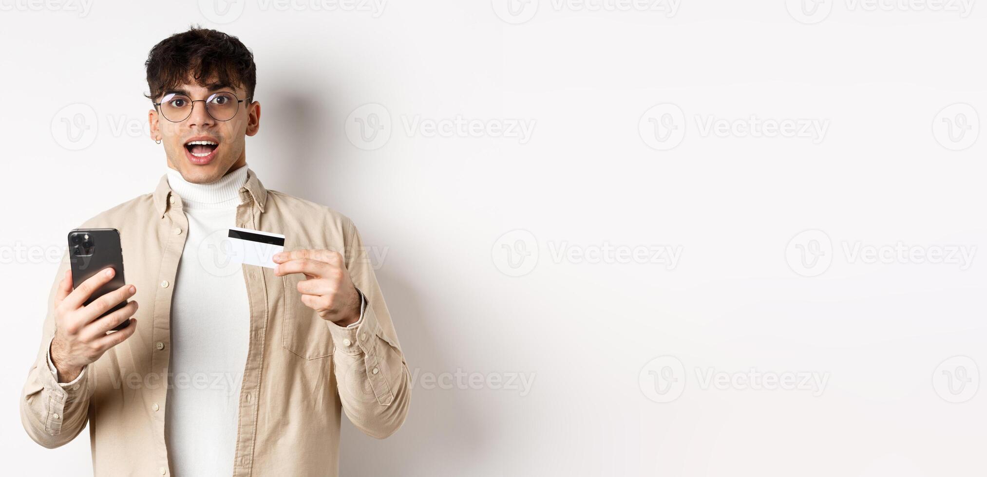 Excited young man shopping online, holding mobile phone and plastic credit card, making purchase in internet, standing on white background photo