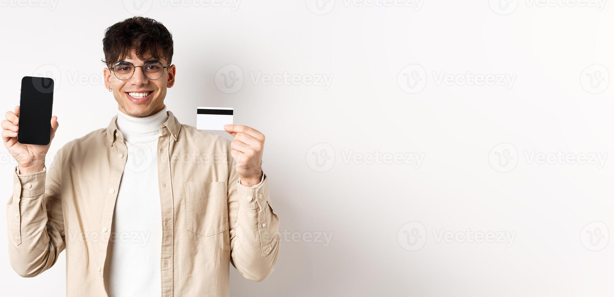 Online shopping. Natural guy in glasses showing empty smartphone screen and plastic credit card, smiling pleased, recommending bank, standing on white background photo