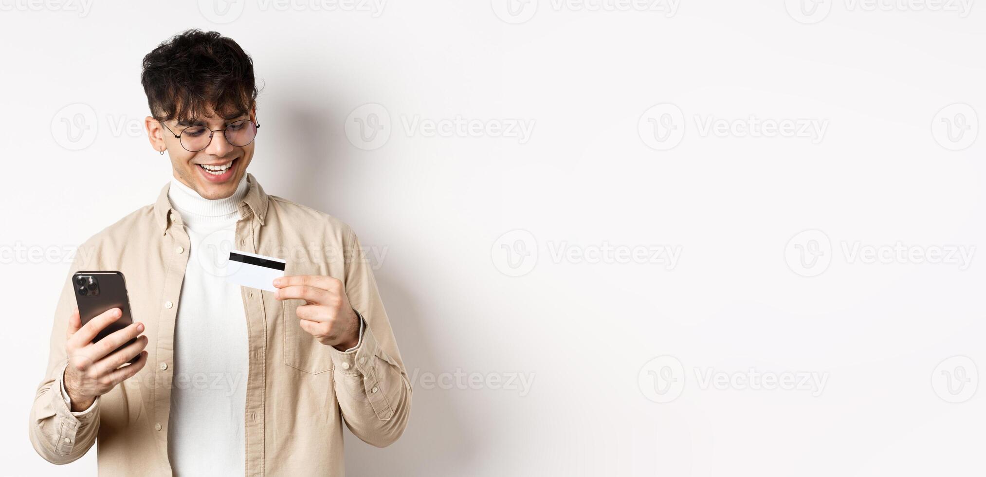 Happy young guy in glasses making online purchase, looking at plastic credit card and holding smartphone, standing on white background photo