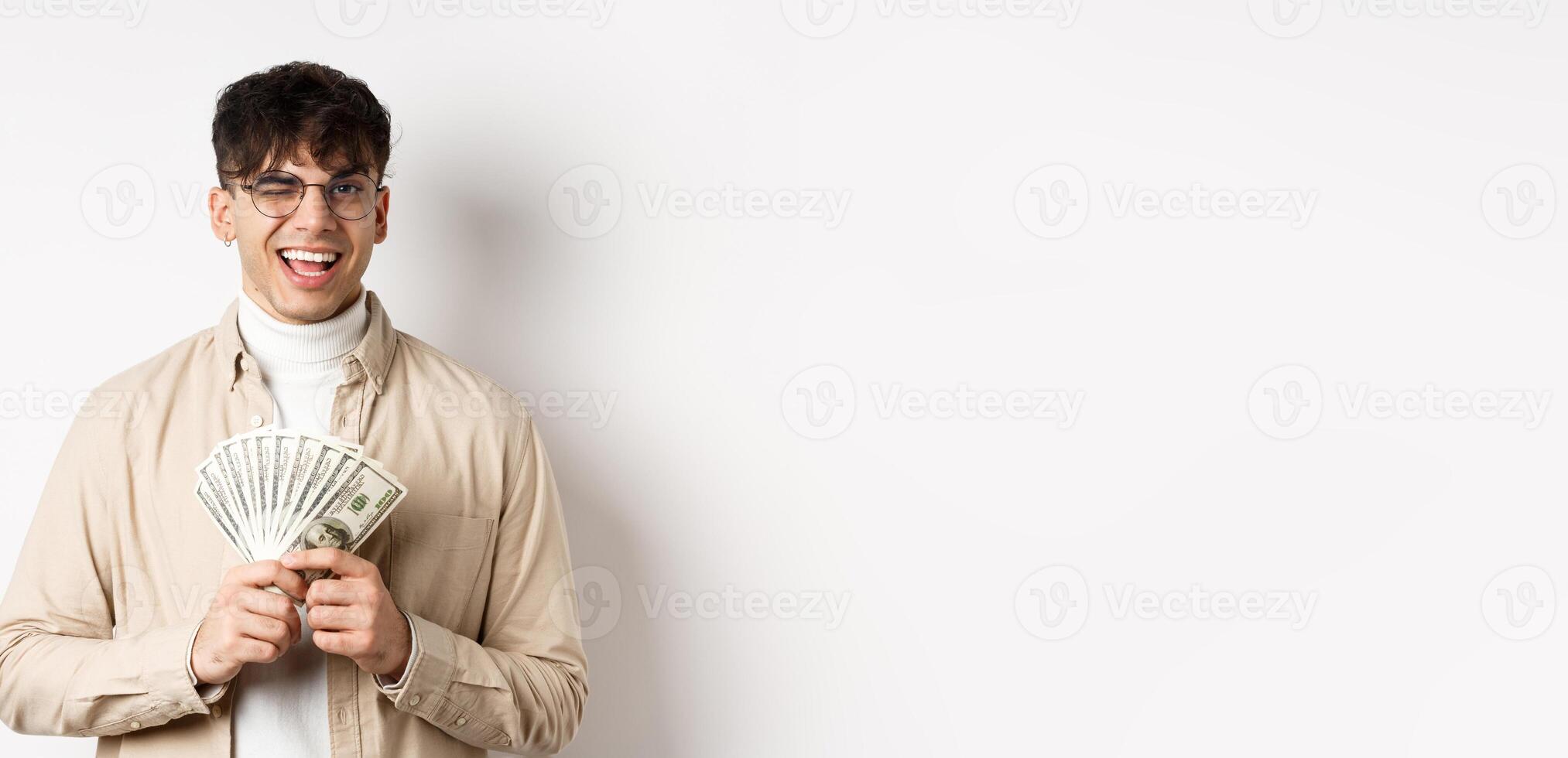 Handsome young man winking and showing dollar bills, holding fan of money banknotes and smiling pleased, making money, standing on white background photo