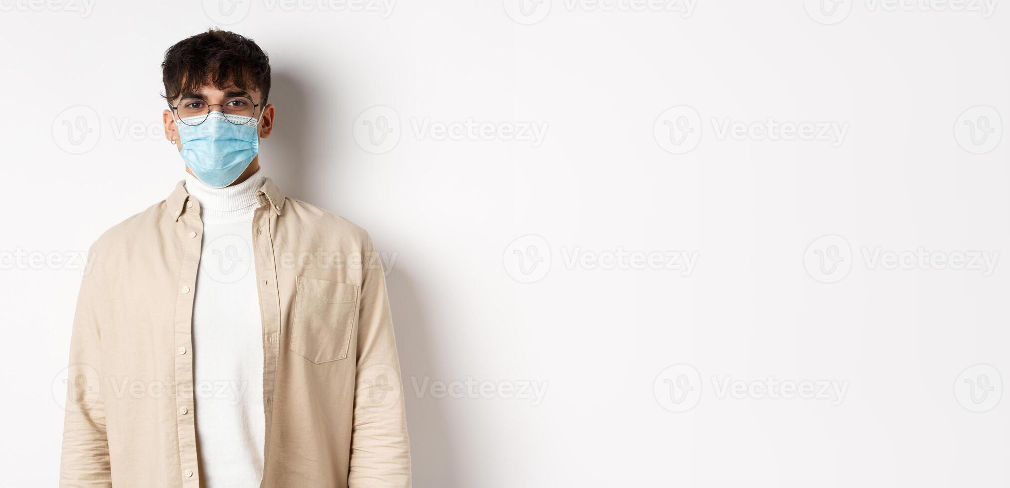 Young man in face mask from covid-19 looking cheerful at camera, standing on white background. Coronavirus, quarantine and social distancing concept photo