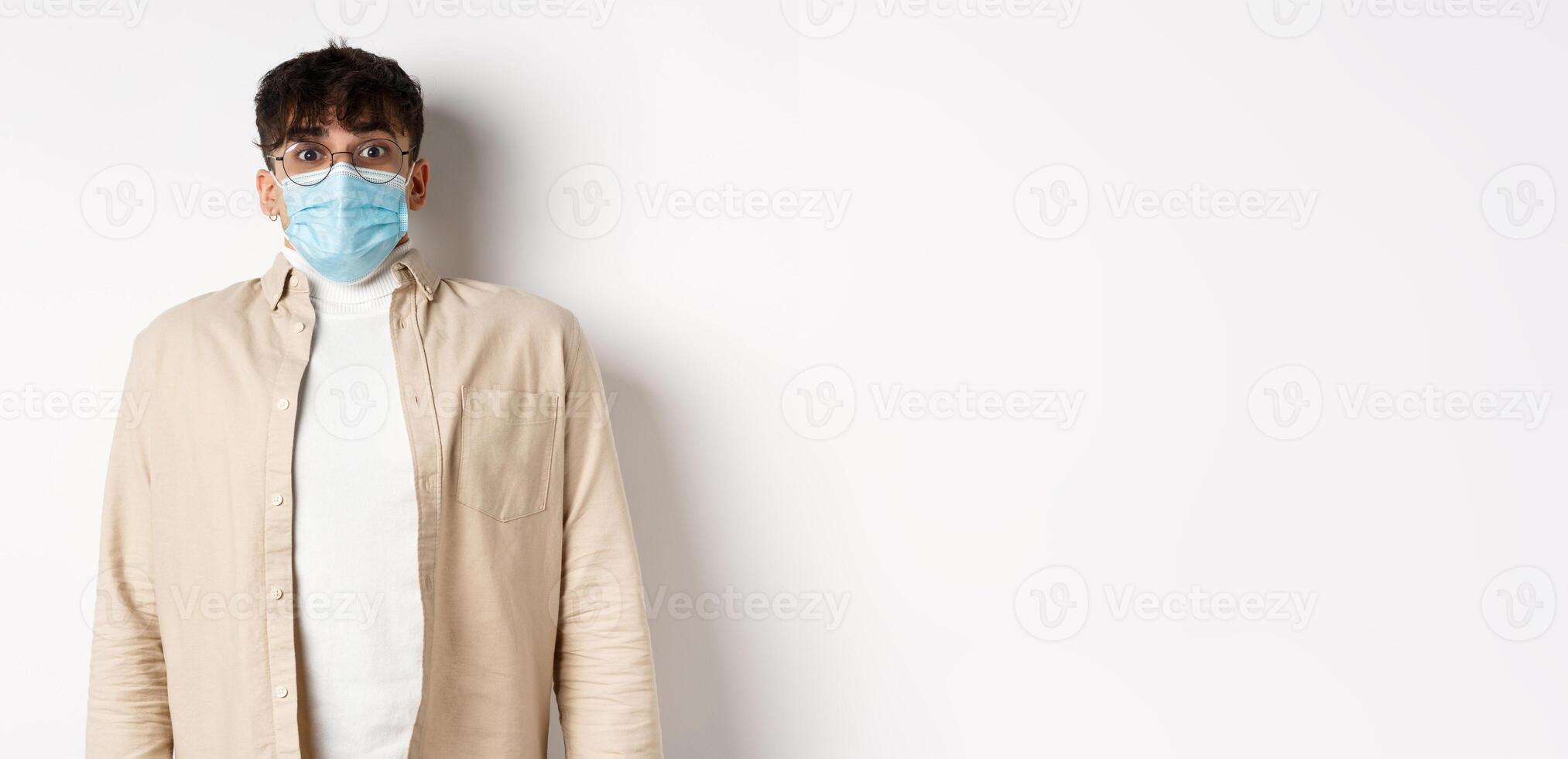 Young man in sterile medical mask from covid-19 looking surprised at camera, standing on white background. Coronavirus, quarantine and social distancing concept photo