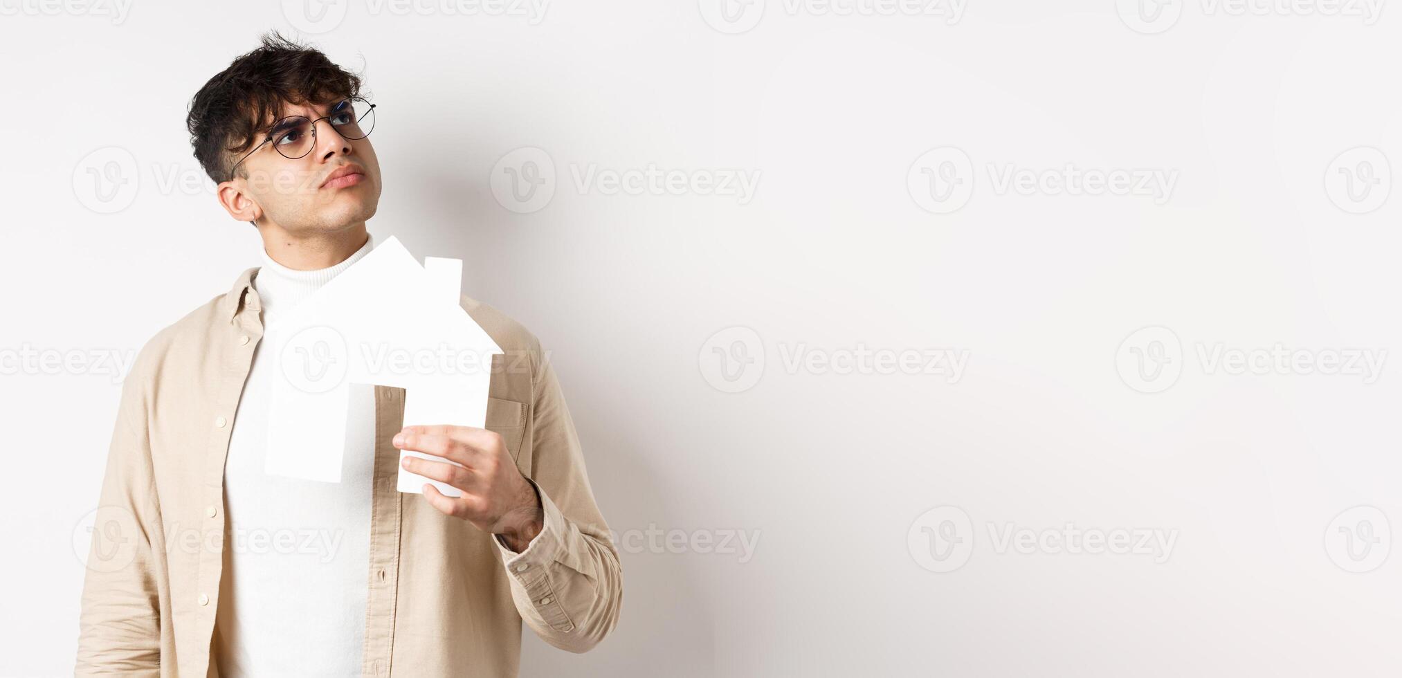 Real estate and mortgage concept. Pensive young man thinking of new apartment, holding paper house cutout and look at upper right corner logo, standing on white background photo