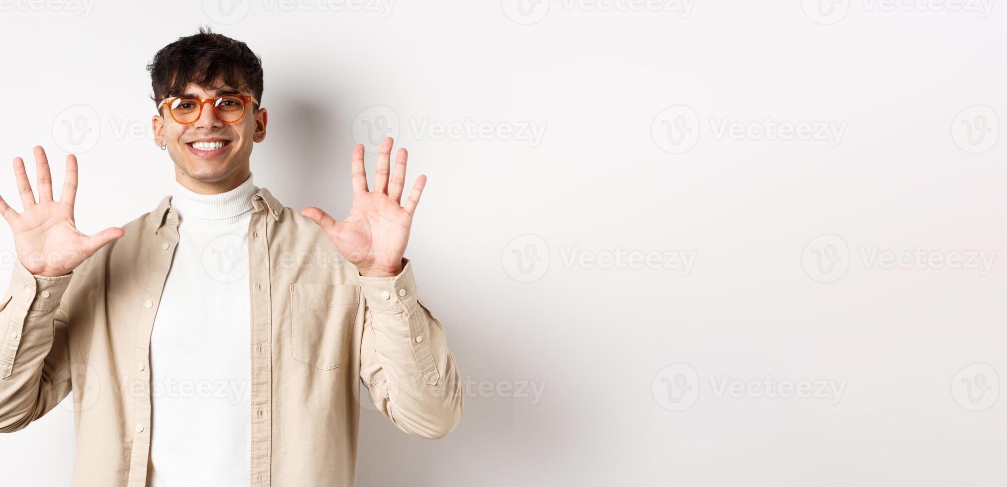 Modern guy in glasses and stylish outfit, showing ten fingers number and smiling, standing on white background photo