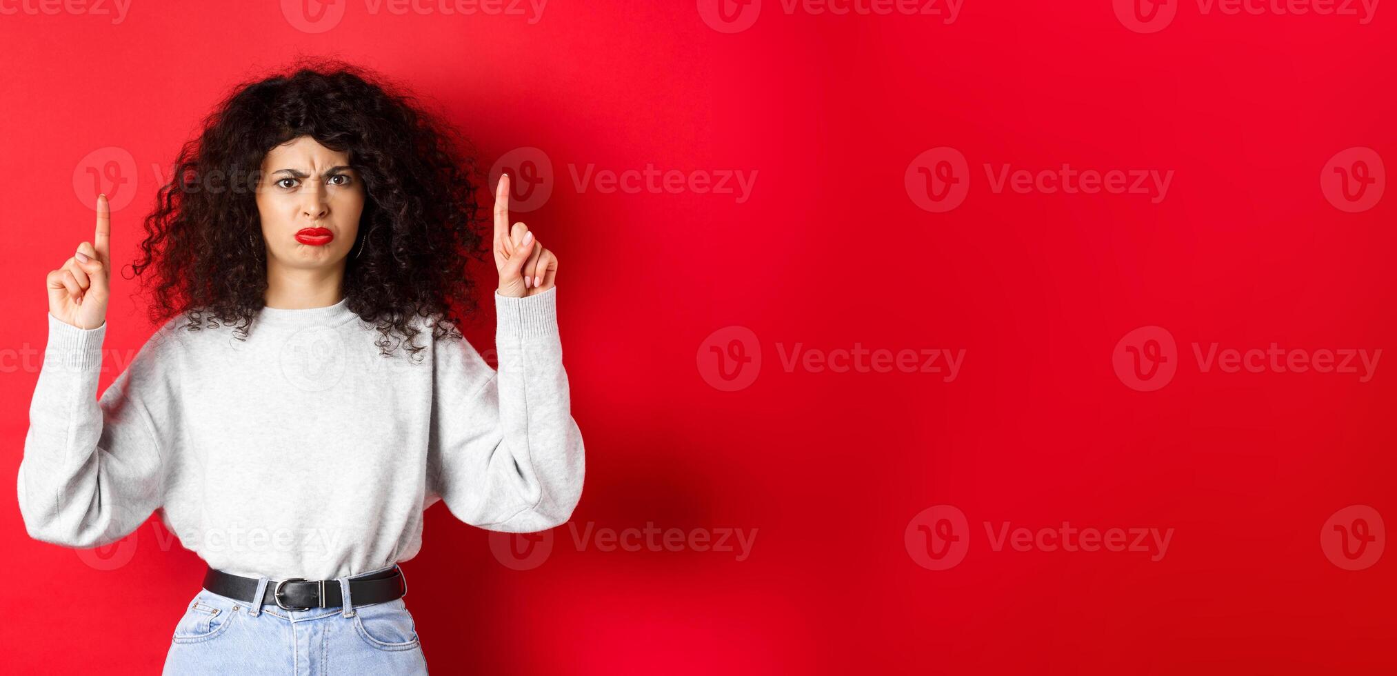 Grumpy young woman with curly hair frowning and grimacing unsatisfied, pointing fingers up at something bad, complaining on company, red background photo