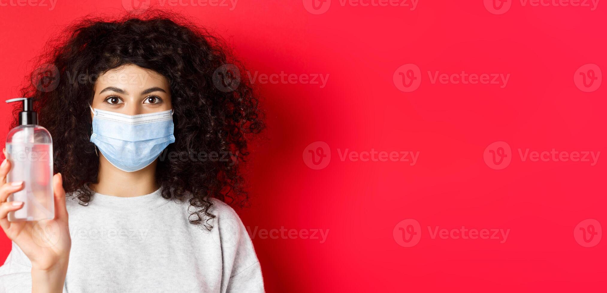 Covid-19, pandemic and quarantine concept. Young woman in medical mask showing bottle of hand sanitizer, demonstrate antiseptic, red background photo