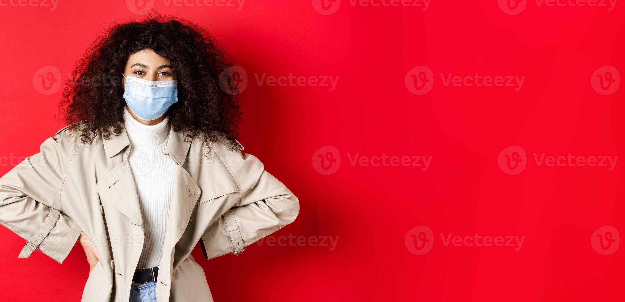 Covid-19, pandemic and quarantine concept. Stylish modern woman with curly hair, wearing trench coat and medical mask, smiling at camera, red background photo