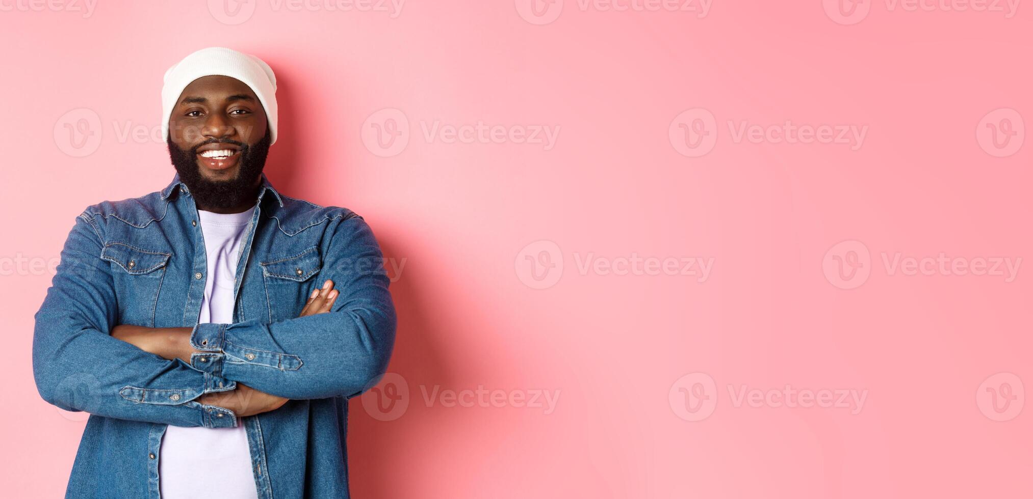 Handsome hip hop style Black man in beanie and denim shirt, smiling confident, cross arms on chest and staring at camera on pink background photo