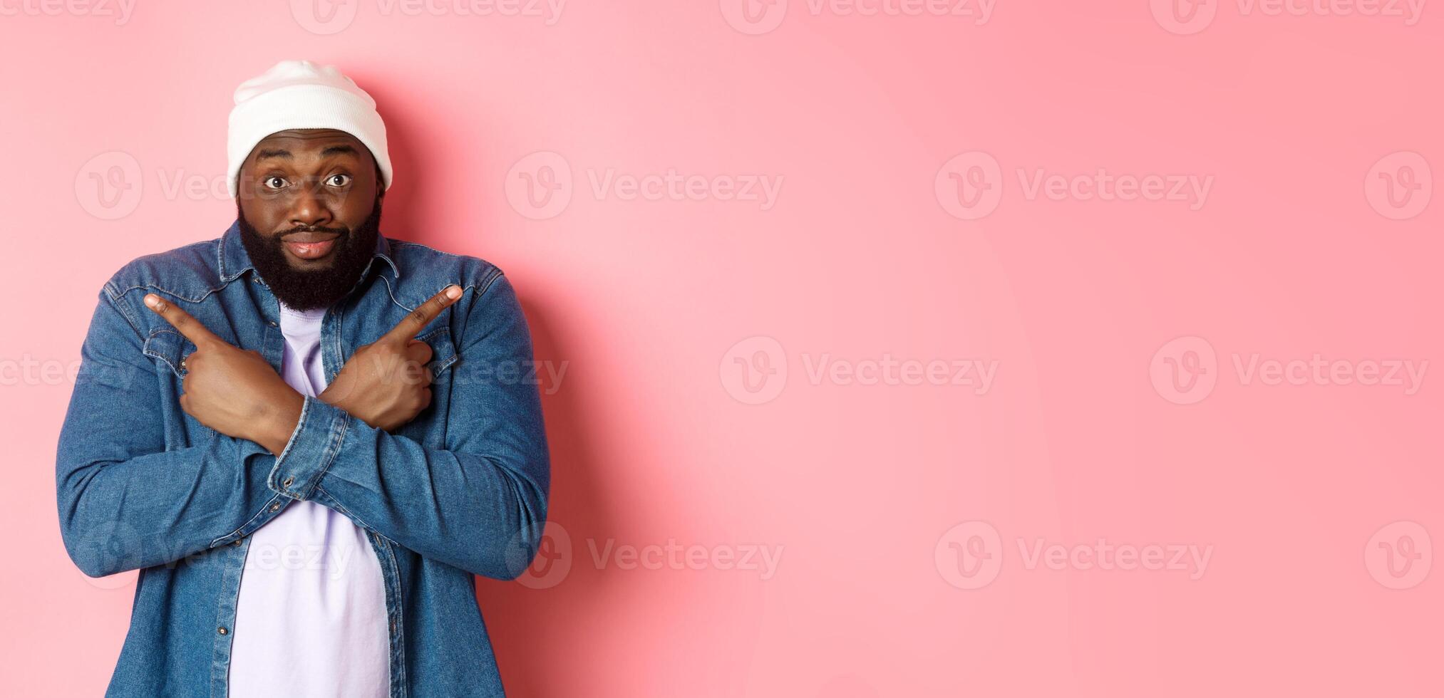 Confused Black man asking help with choice, pointing sideways and staring at camera, making decision, standing over pink background photo