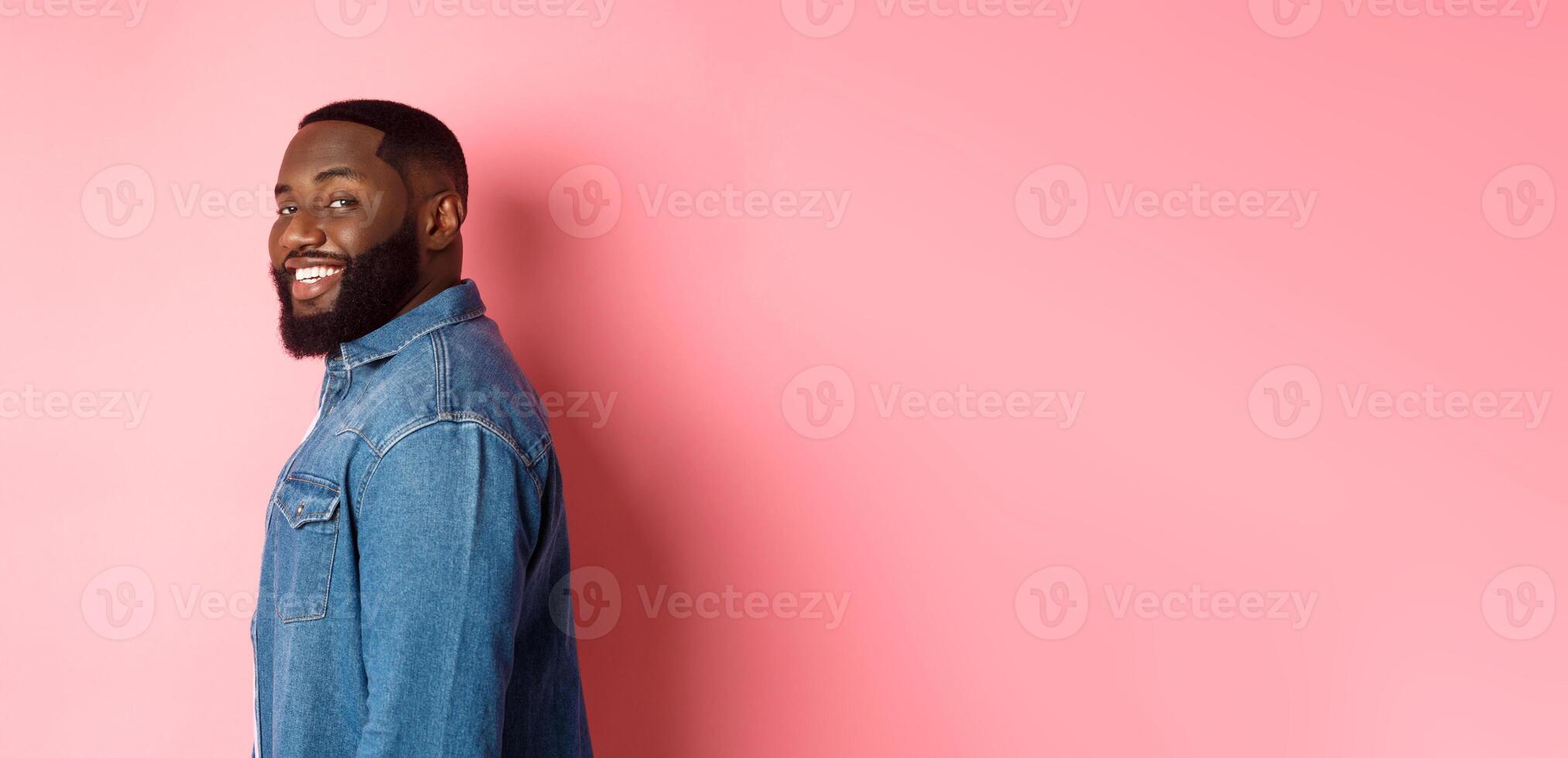 Handsome african-american man with beard, turn face at camera and smiling confident, standing over pink background photo