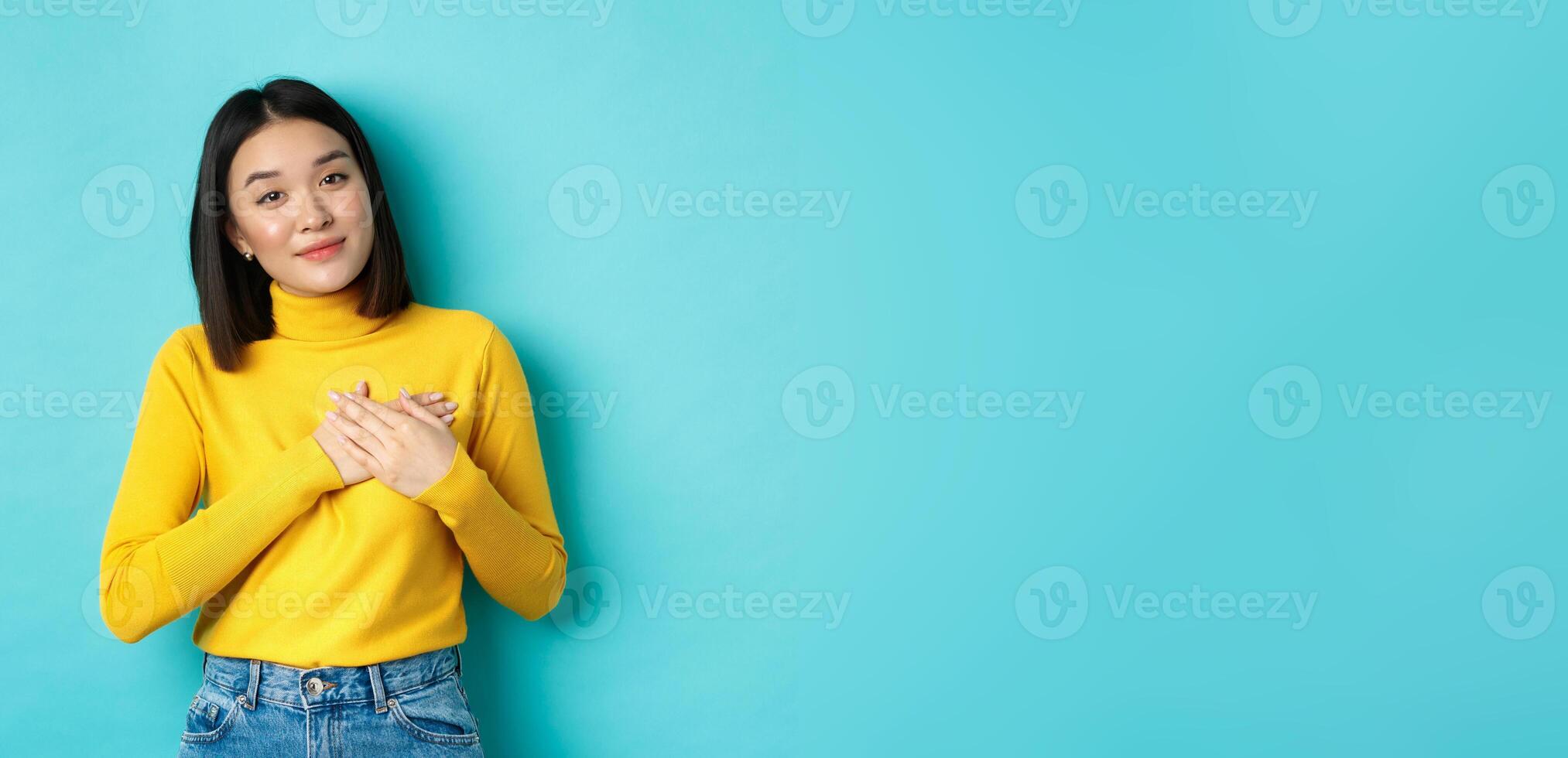 Portrait of beautiful heartfelt woman holding hands on heart, smiling and listening compassionate, standing over blue background in yellow pullover photo