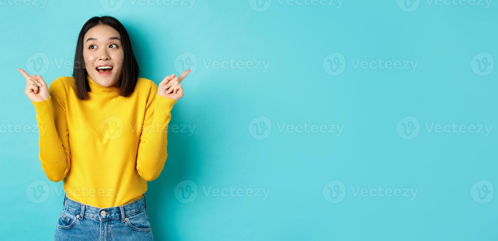 Beauty and fashion concept. Excited asian girl in yellow pullover making choice, pointing fingers sideways and choosing between two options, standing against blue background photo