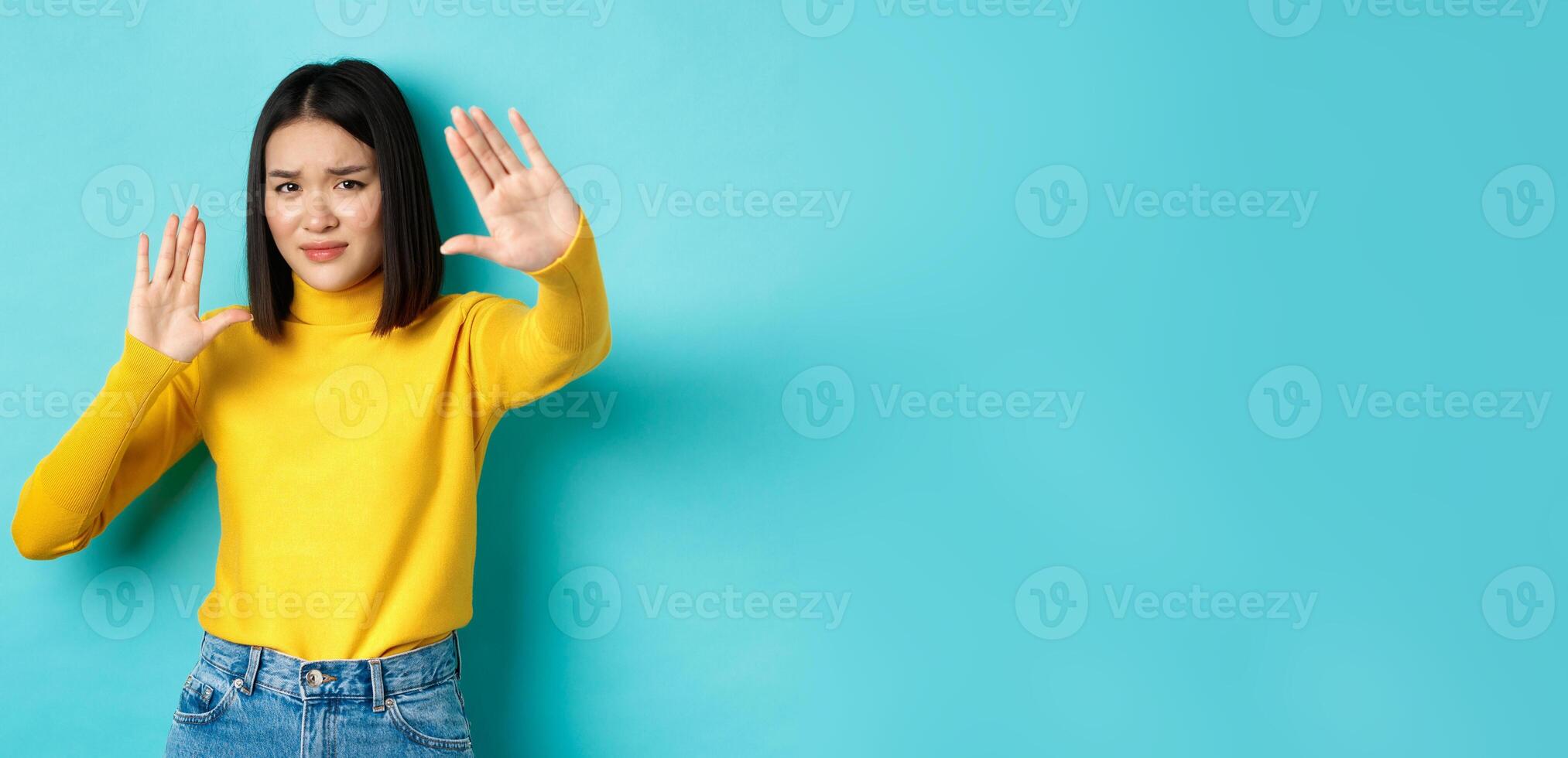 Image of woman feeling uncomfortable in front of camera, cover face with hands and frowning, begging to stop, standing against blue background photo