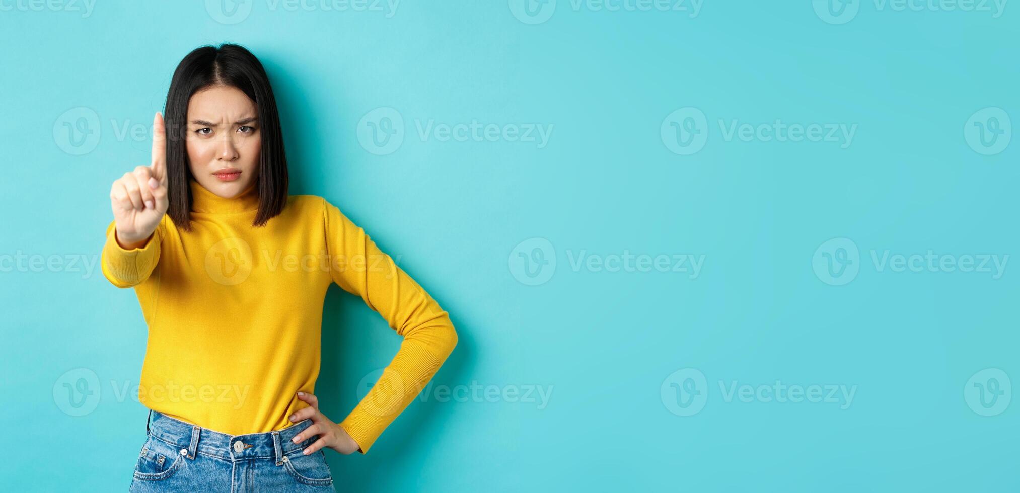 Confident and serious woman tell no, showing extended finger to stop and prohibit something bad, frowning and looking at camera self-assured, standing over blue background photo
