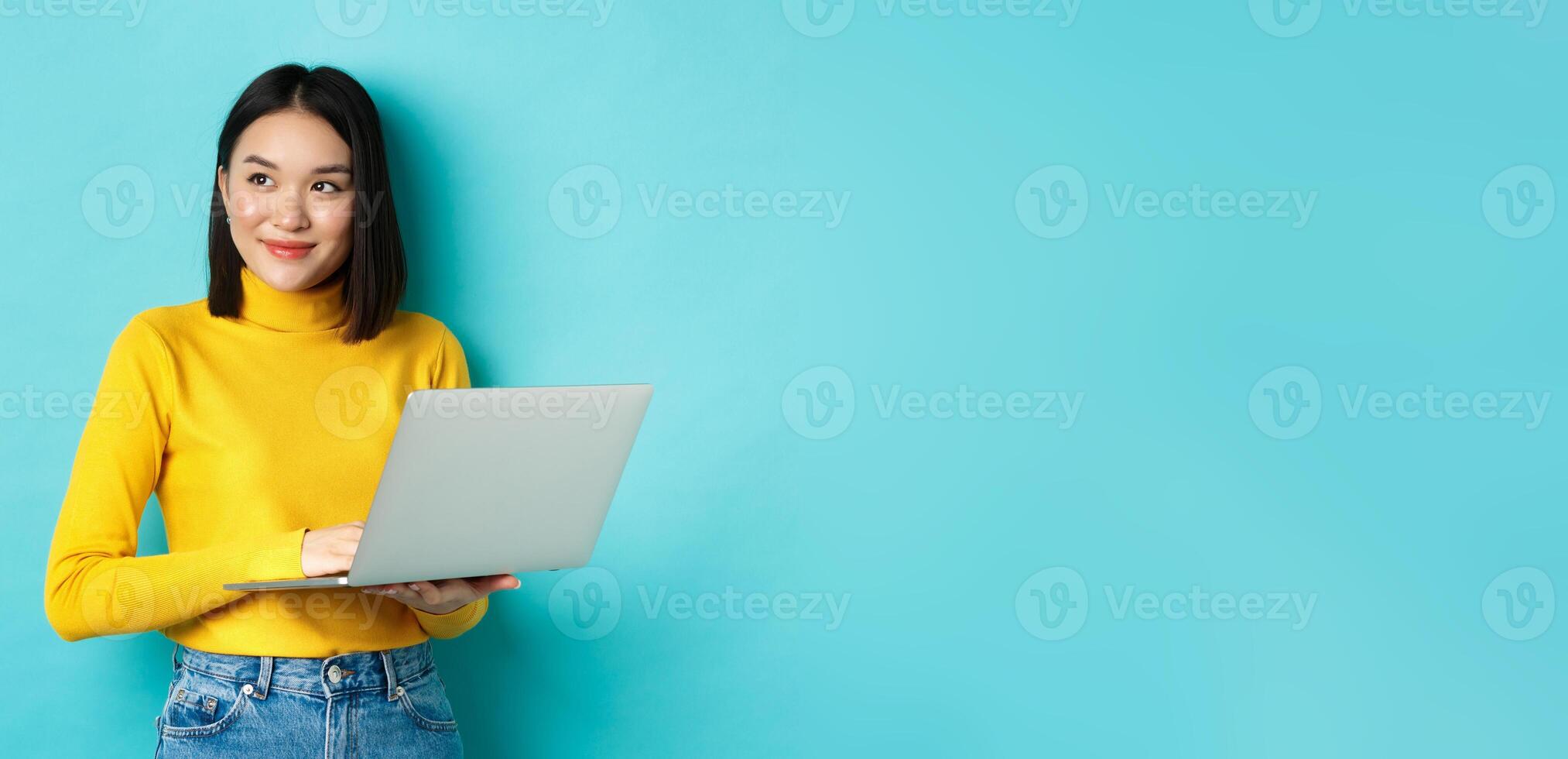 Image of stylish korean woman working on laptop and looking away with dreamy smile, standing with computer against blue background photo