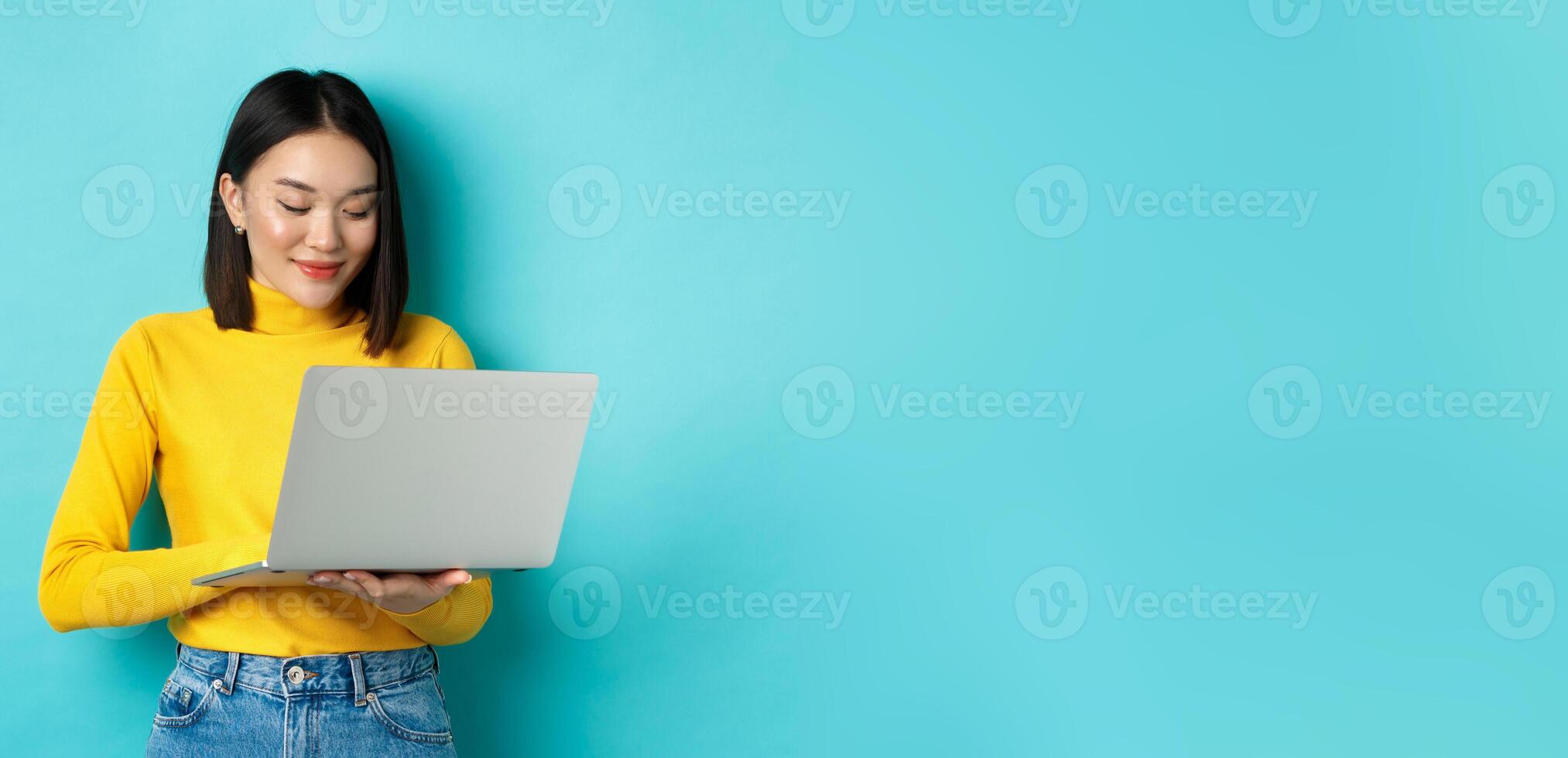 Beautiful asian female student working on laptop, typing on keyboard and looking at screen with pleased smile, standing over blue background photo