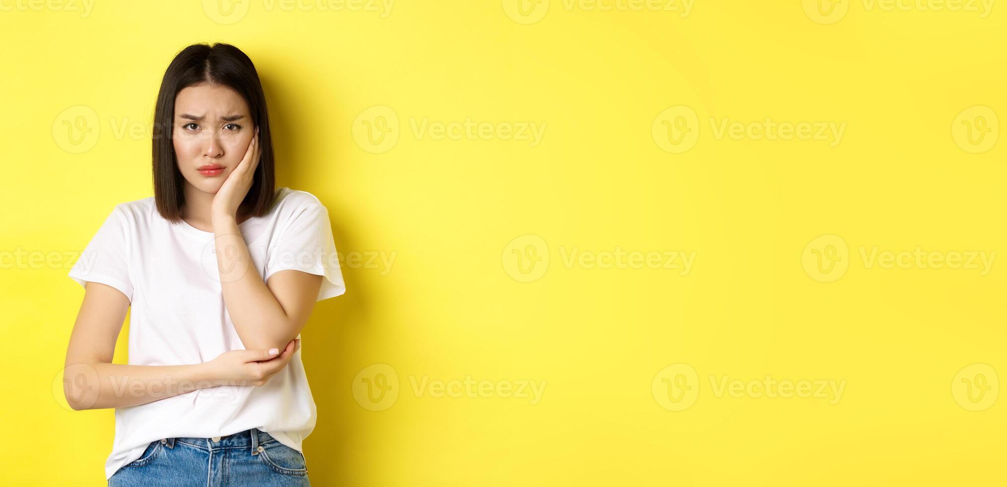 Sad asian woman touching cheek and frowning, having toothache, need see dentist, standing gloomy against yellow background photo