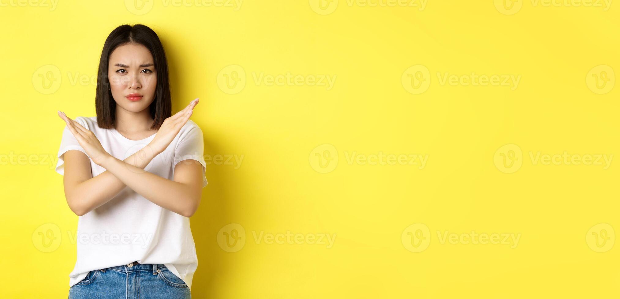 Serious and assertive korean woman showing cross stop gesture, frowning and telling no, prohibit action, disapprove something bad, standing over yellow background photo