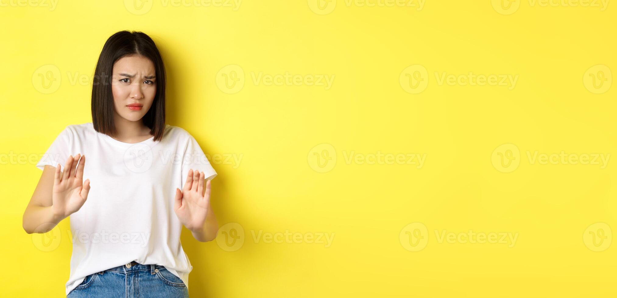 Please no. Asian woman, victim of assualt or domestic abuse, pleading with hands raised up in defense, frowning sad, begging stop, standing over yellow background photo