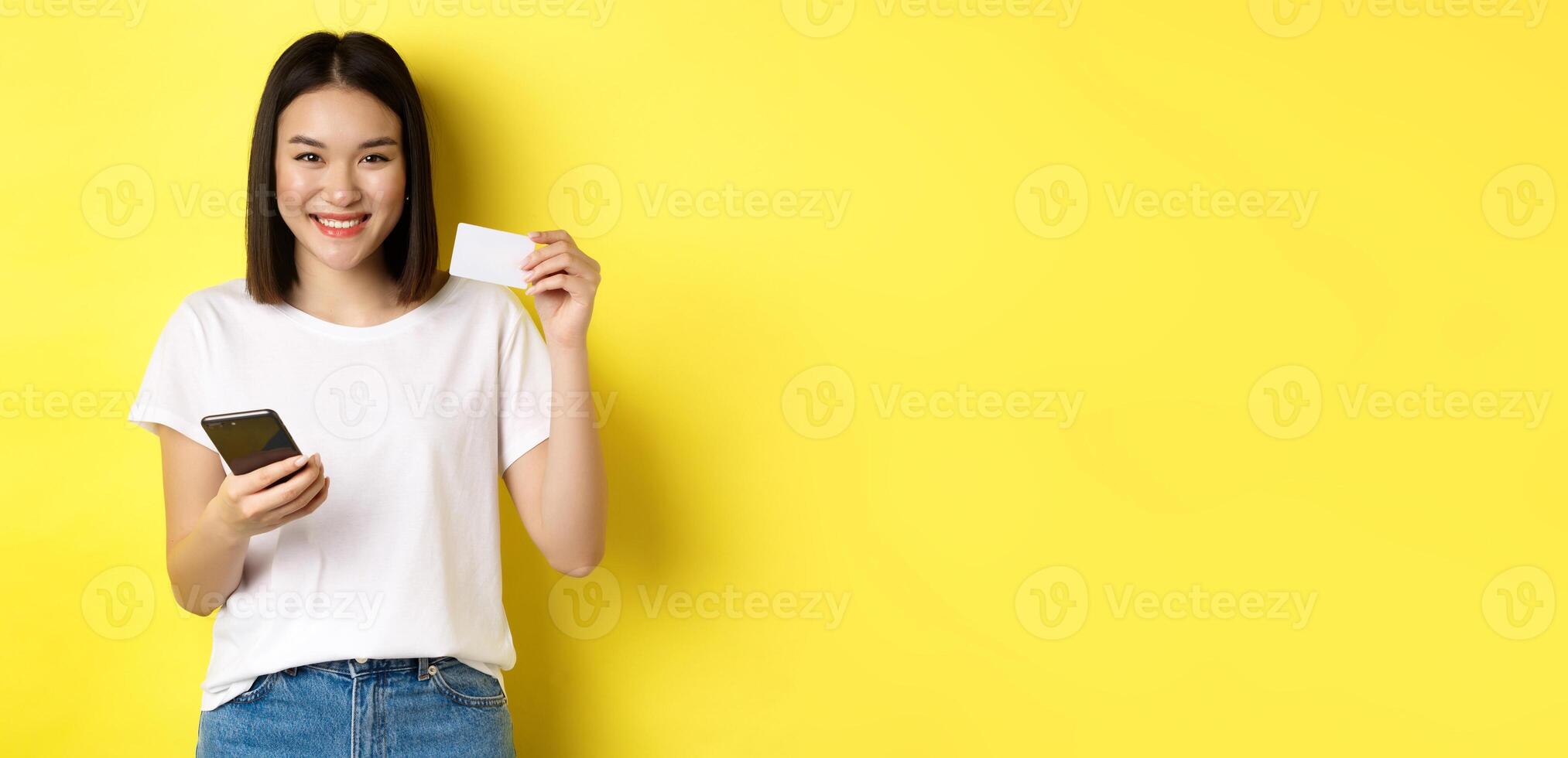 Attractive korean woman paying online with smartphone, showing plastic credit card and smiling, standing over yellow background photo