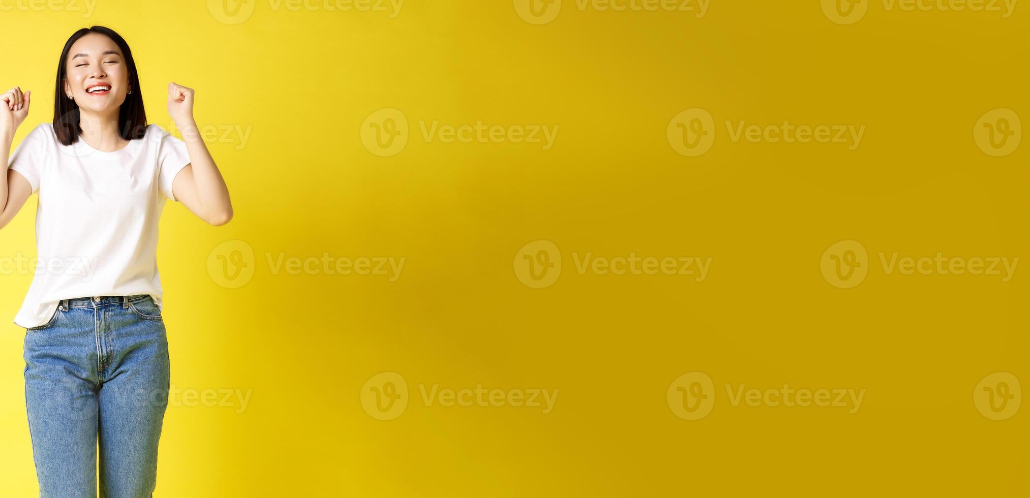 Full size shot of cheerful asian woman in jeans and white t-shirt jumping from happiness, winning prize and rejoicing, celebrating and shouting yes with joy, yellow background photo