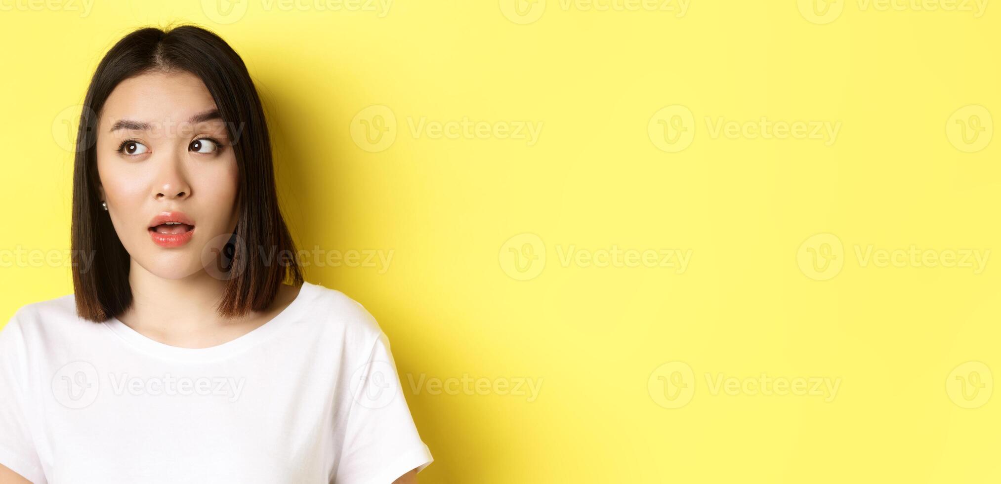 Close up of beautiful asian woman with clean perfect skin, raising eyebrow and looking left with intrigued face, standing over yellow background photo