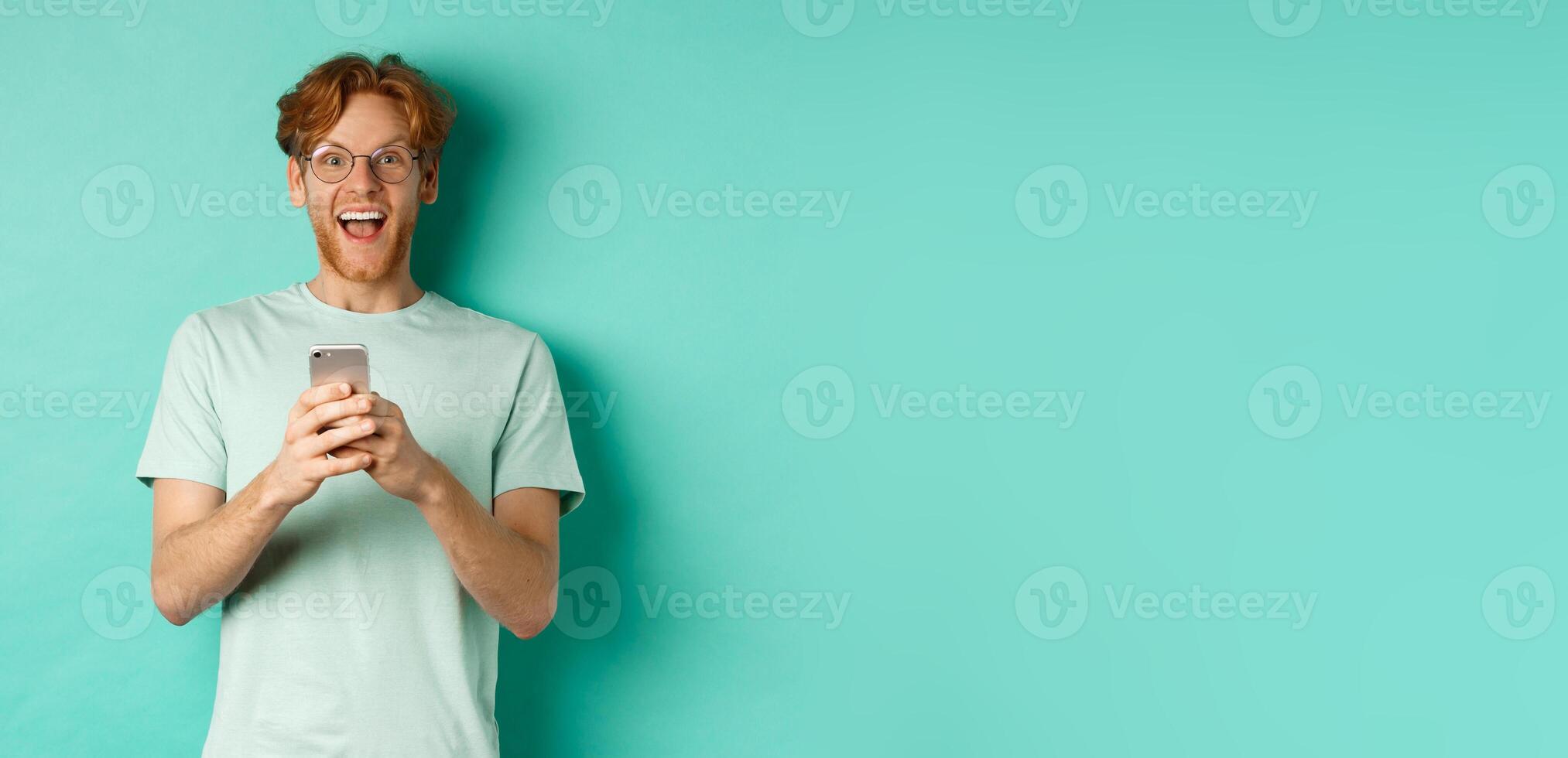 Surprised redhead man in glasses looking amazed at camera after reading promo offer on smartphone, standing against turquoise background photo