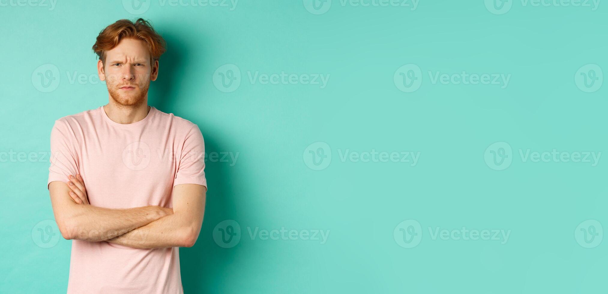 Young man with ginger hair and beard looking offended, feeling insulted and staring at camera, sulking at you, cross arms on chest defensive, standing over turquoise background photo