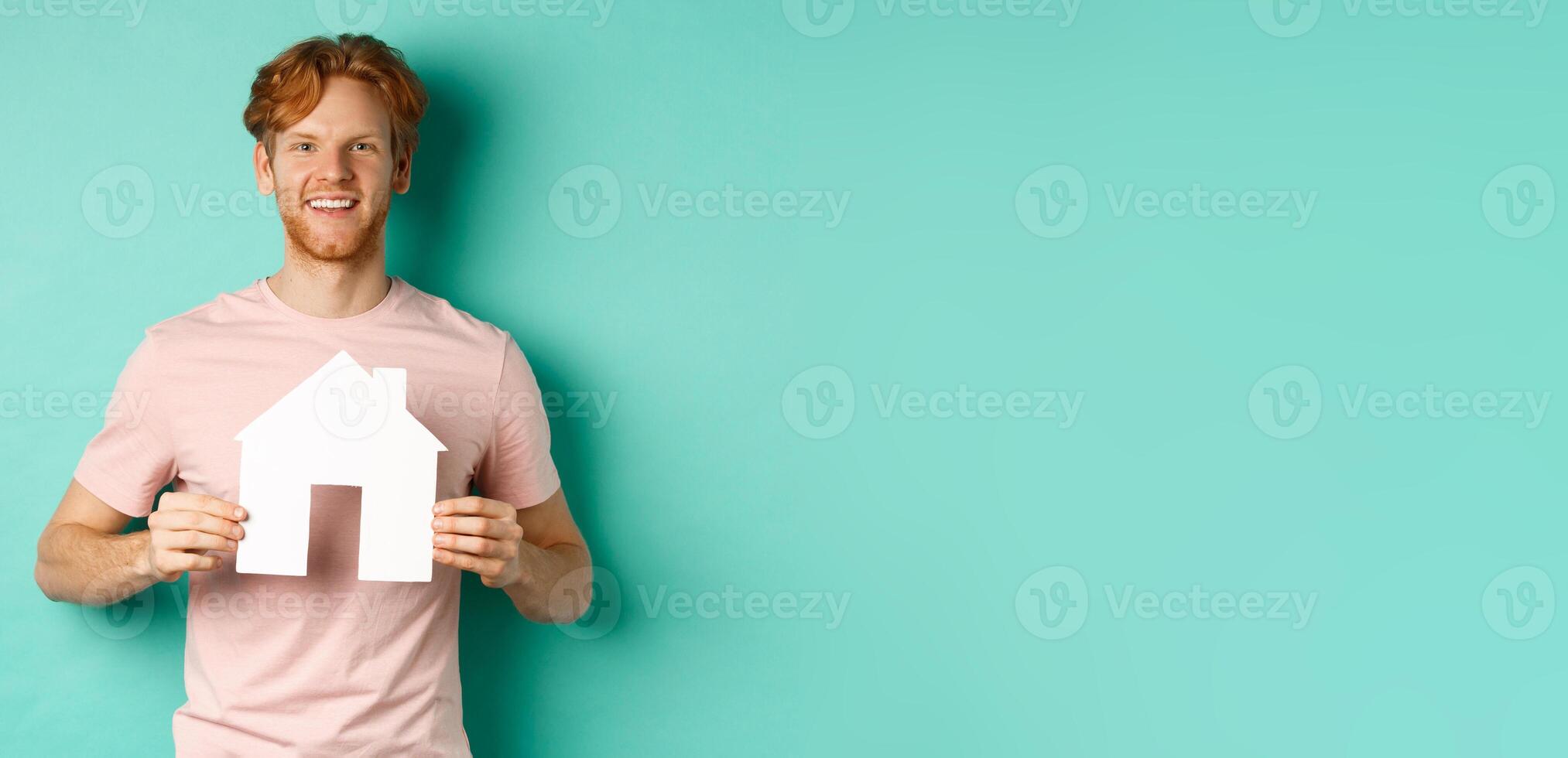 Real estate concept. Young man with red hair, wearing t-shirt, showing paper house cutout and smiling happy, standing over mint background photo
