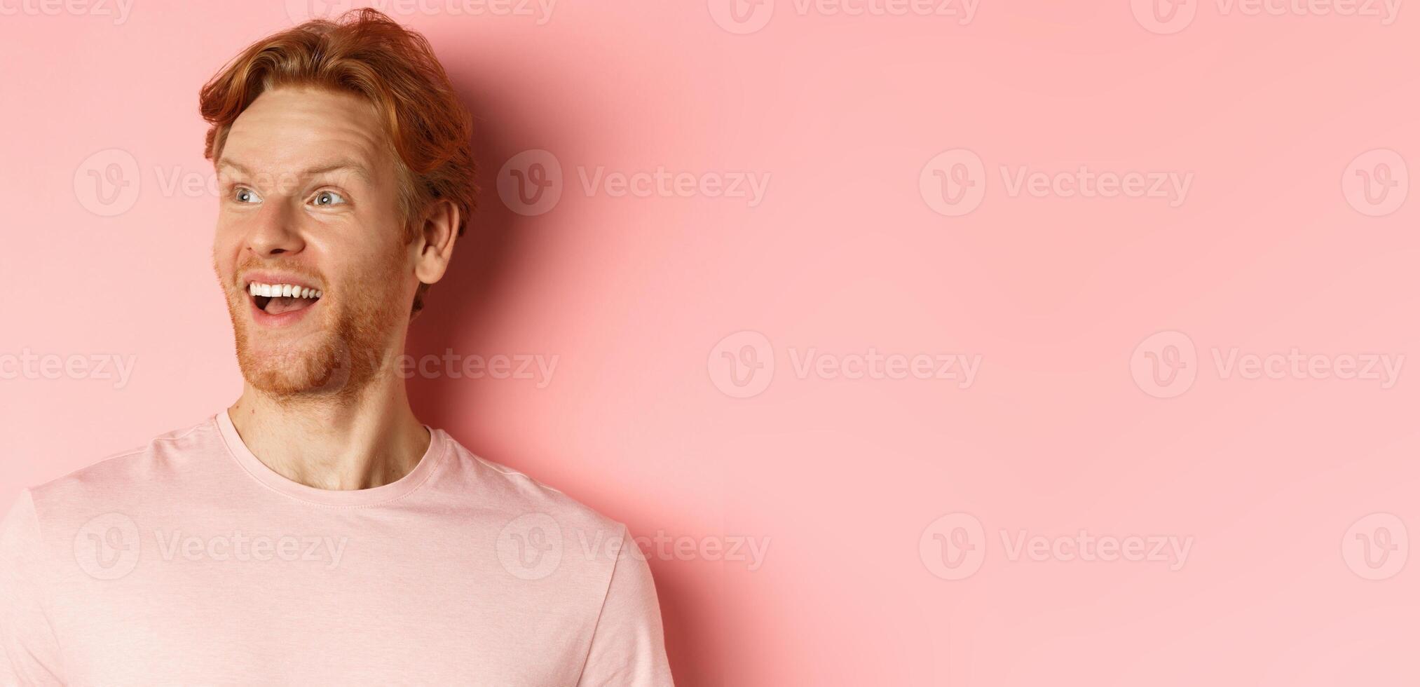 Close-up of handsome caucasian guy with red hair and beard, looking left with happy smile, checking out promo offer, standing over pink background photo