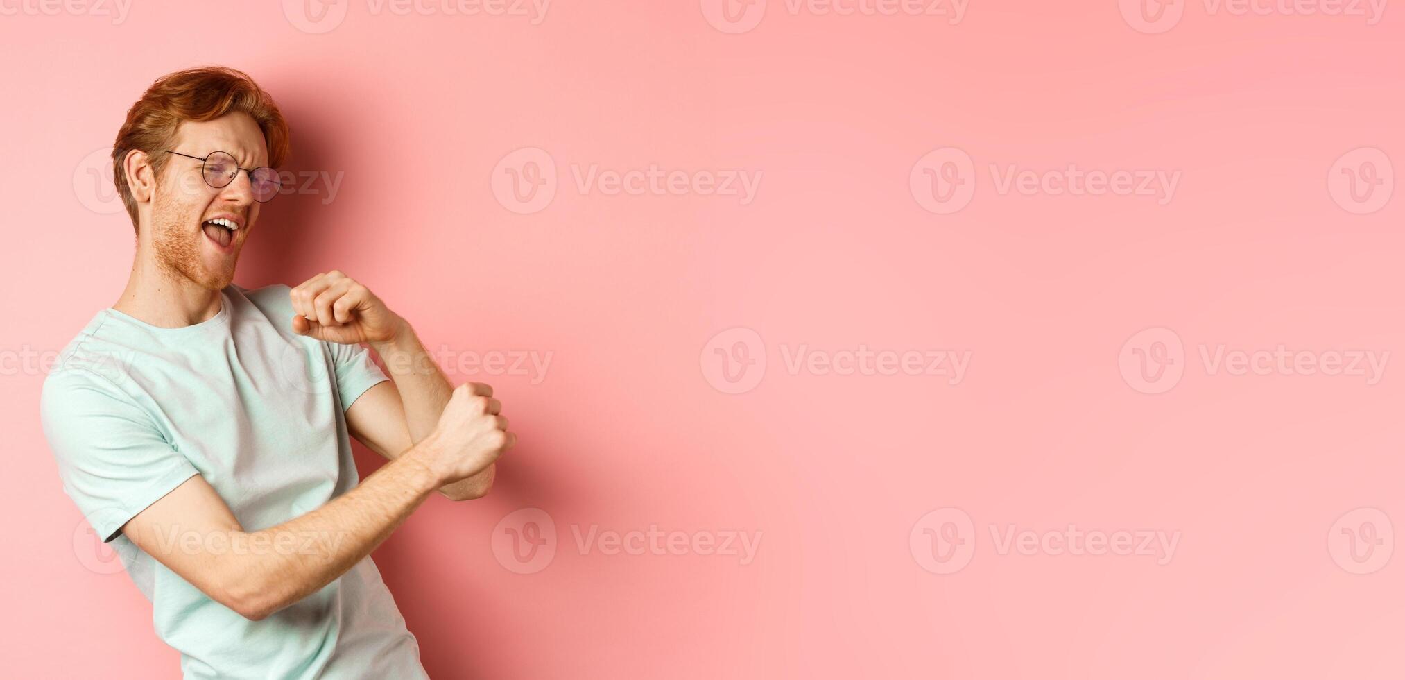 Summer vacation concept. Happy young man with red hair dancing and having fun, singing along music, standing over pink background photo