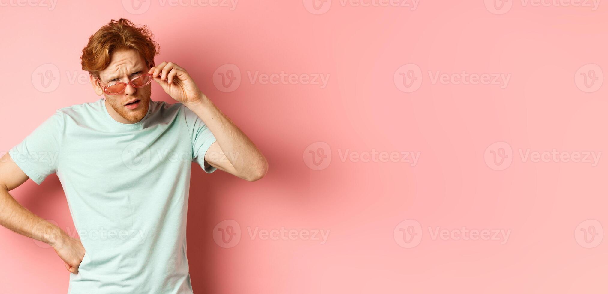 Tourism and vacation concept. Confused funny guy with red messy hair and beard, take-off glasses and frowning puzzled, standing over pink background photo