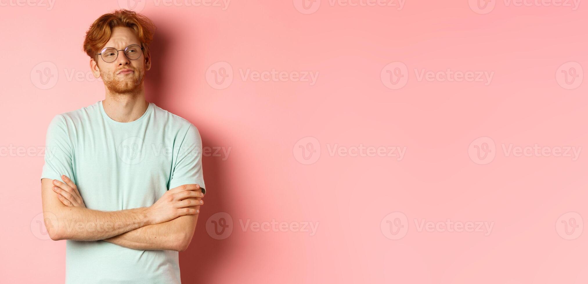 Arrogant redhead guy in glasses cross arms on chest, looking at something with skeptical face, standing over pink background photo