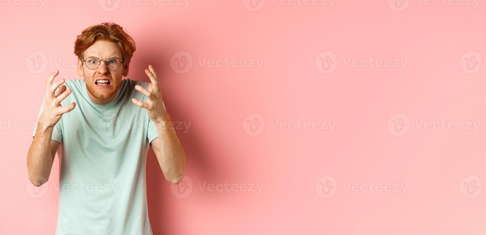 Angry redhead guy in glasses shouting, frowning and shaking hands with frustrated and outraged face, standing over pink background photo