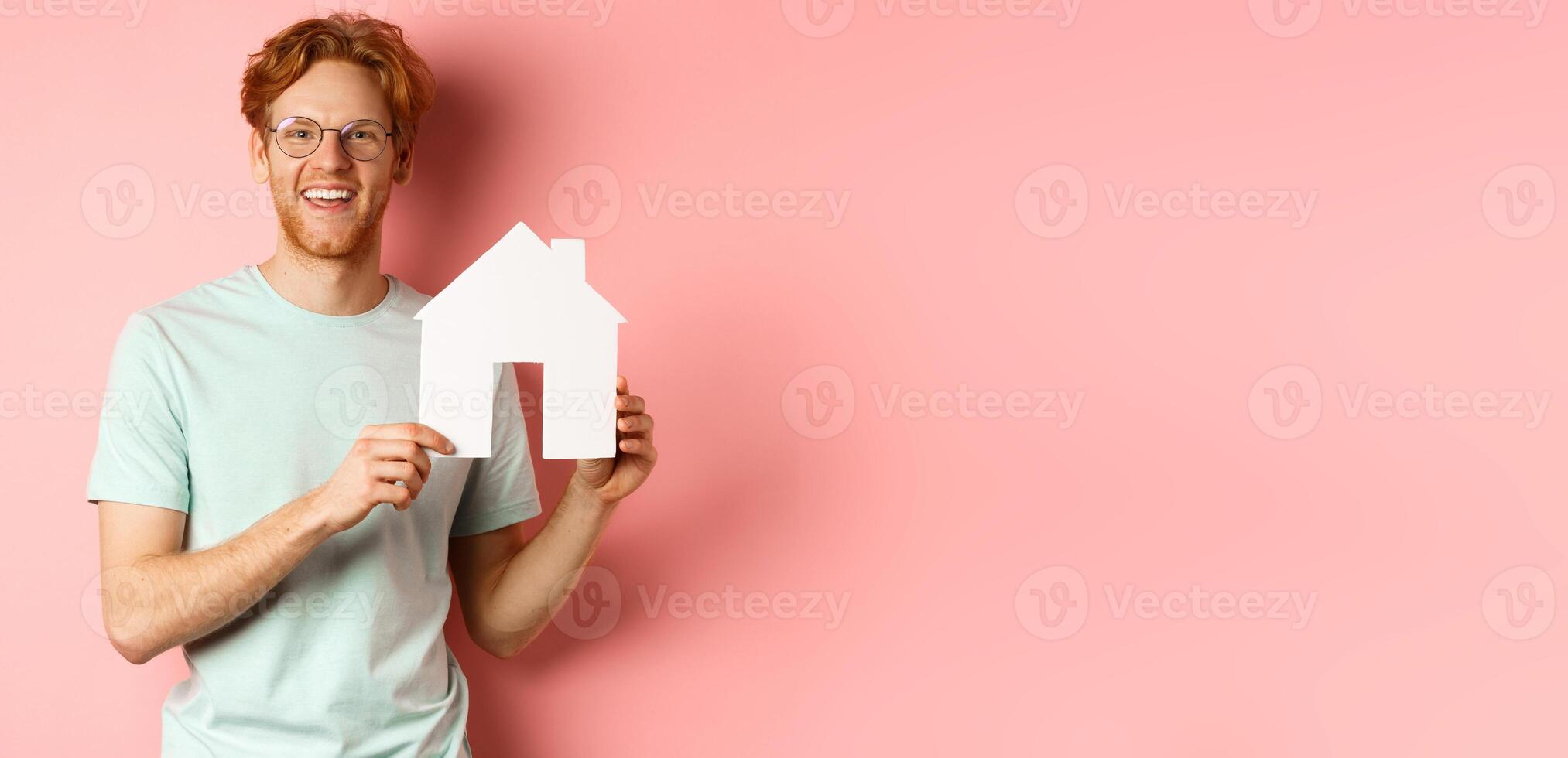 Real estate. Handsome redhead man in t-shirt and glasses, showing paper house cutout and smiling, standing over pink background photo