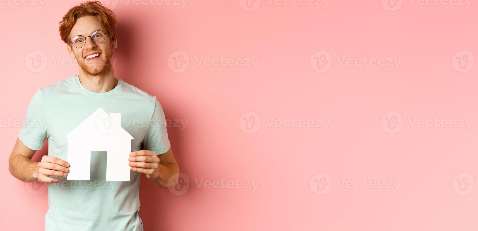 Real estate. Cheerful young man with red hair, wearing glasses and t-shirt, showing paper house cutout and smiling, buying apartment, pink background photo