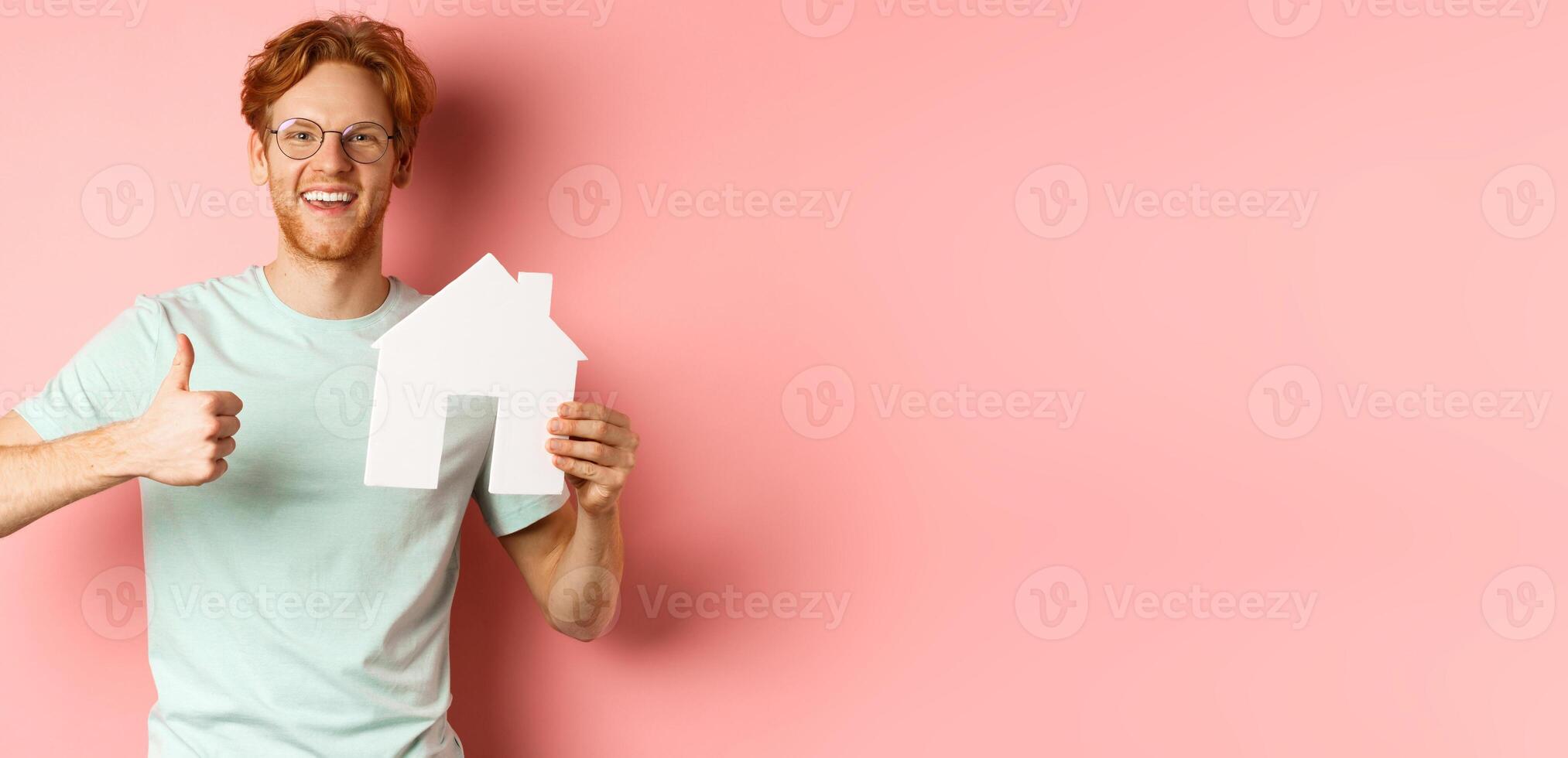 Real estate. Cheerful man in glasses and t-shirt recommending broker agency, showing paper house cutout and thumbs-up, standing over pink background photo