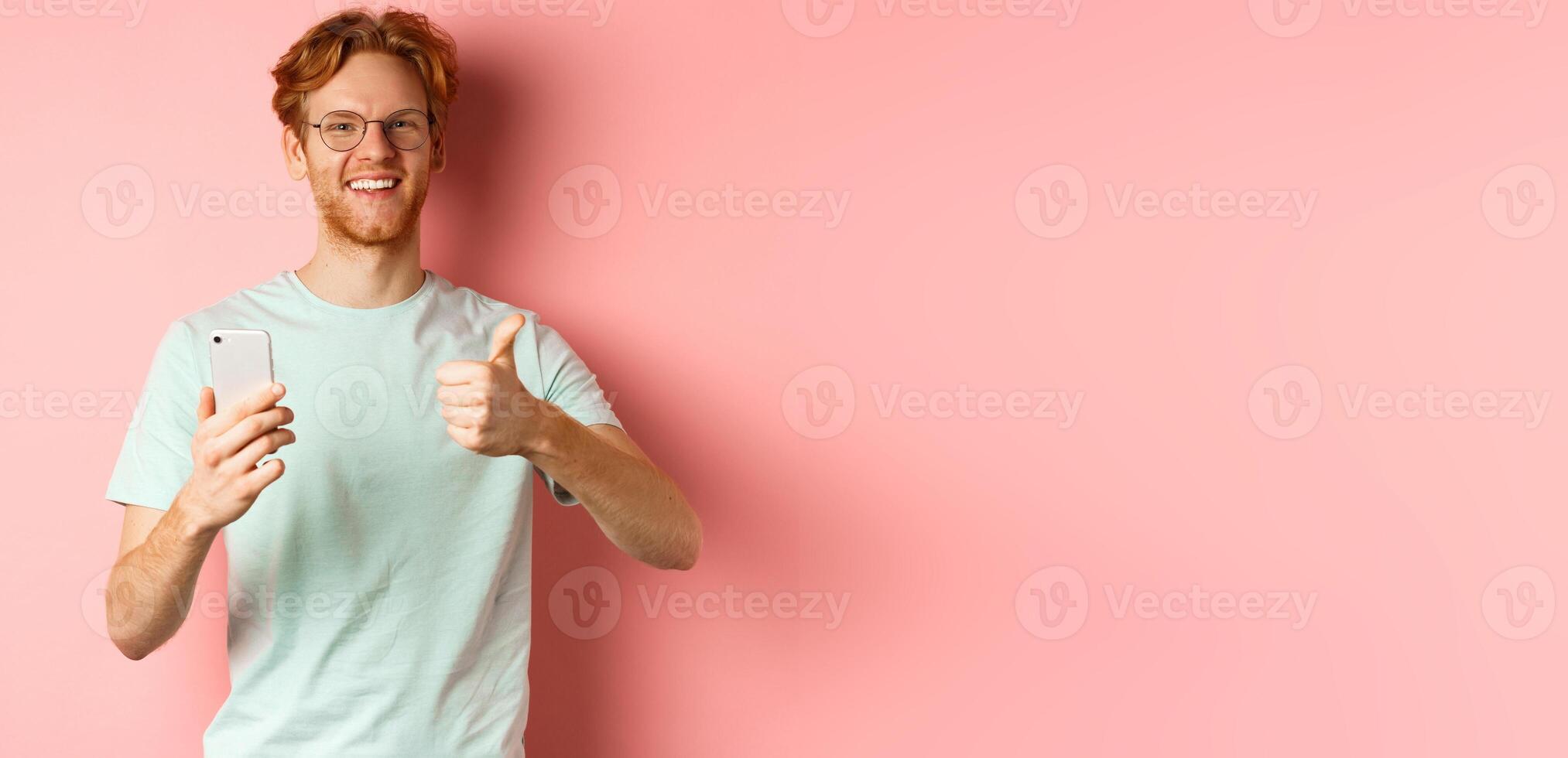 Happy young man with red messy hair, showing thumbs-up while using mobile phone, smiling and praising application, standing over pink background photo