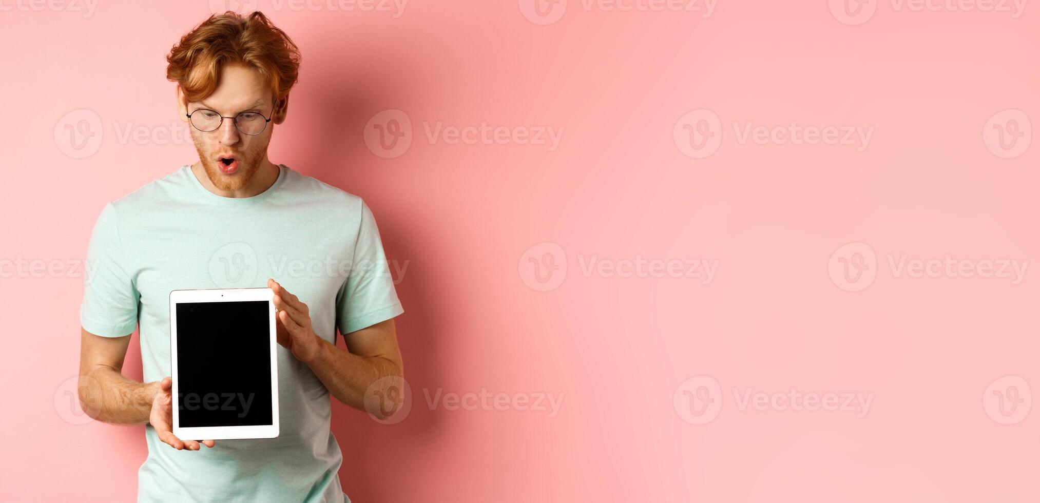 Impressed redhead man in glasses, showing blank digital tablet screen and looking in awe at display, saying wow, standing in t-shirt against pink background photo