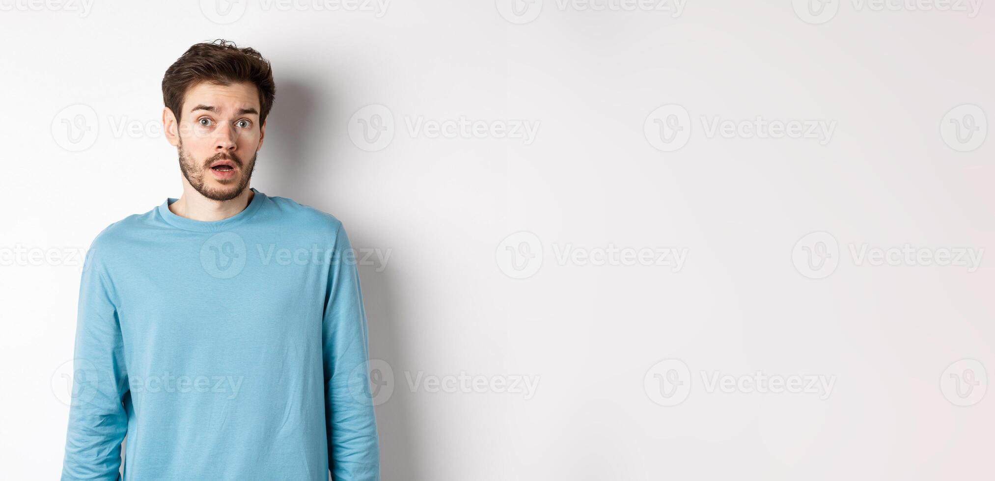 Confused caucasian guy drop jaw and stare startled at camera, standing over white background photo