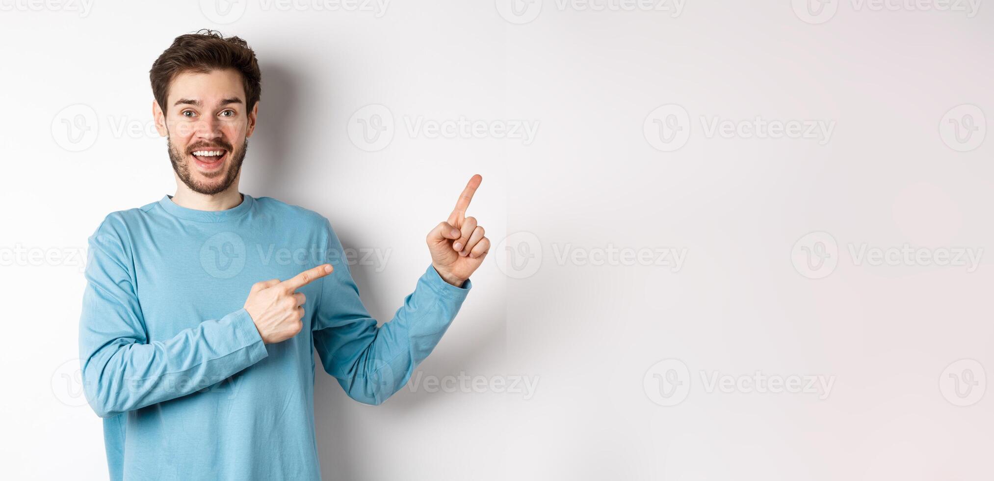Cheerful smiling man pointing fingers right and showing banner or logo on white space for advertisements, recommending product, standing over studio background photo