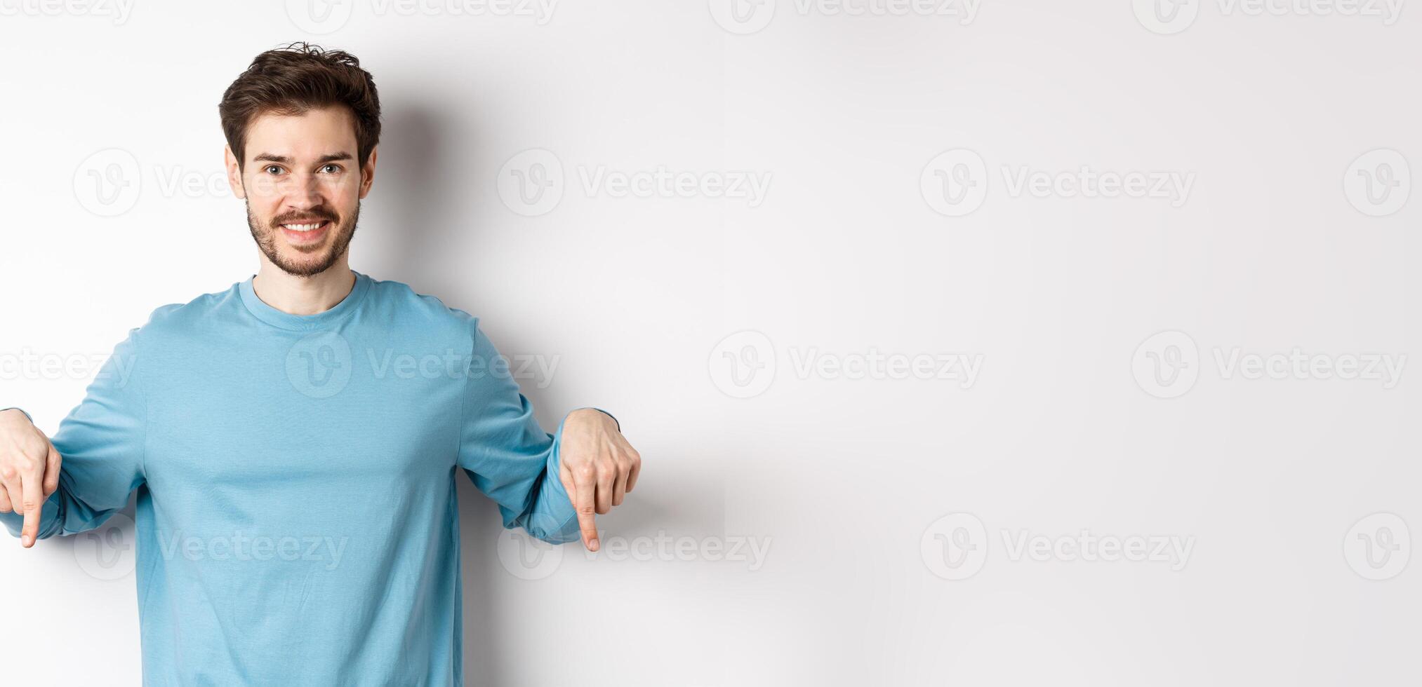 Smiling confident man pointing fingers down, showing promo banner or logo on white background, standing in casual blue sweatshirt photo