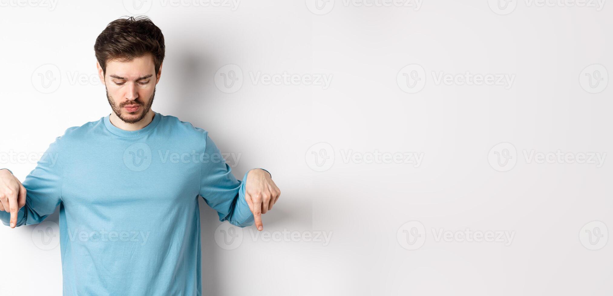 Handsome caucasian man with beard, wearing blue shirt, looking and pointing down at interesting logo banner, standing on white background photo