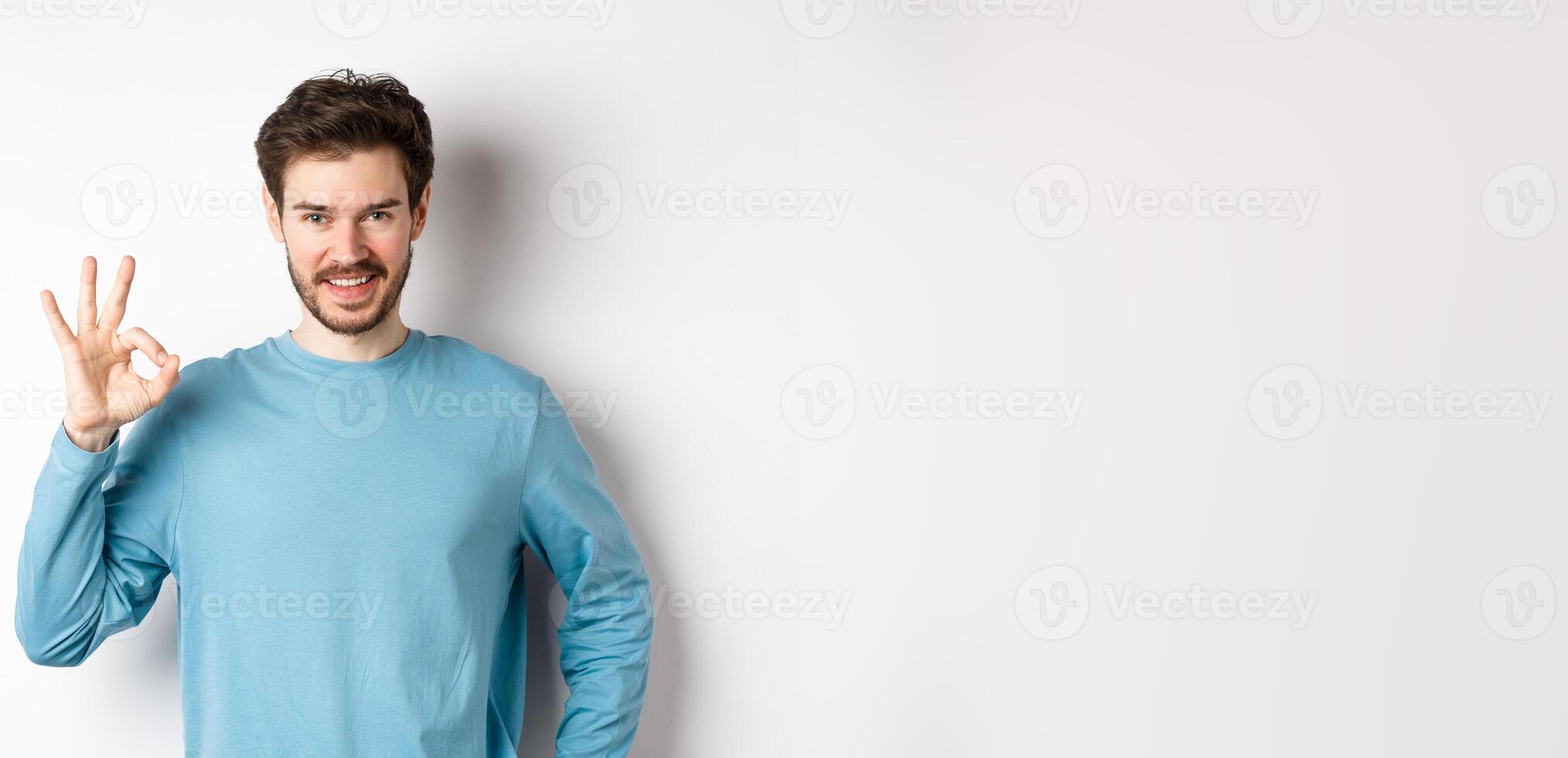 Okay. Handsome smiling man showing ok sign and looking confident, recommend or praise something good, standing over white background photo