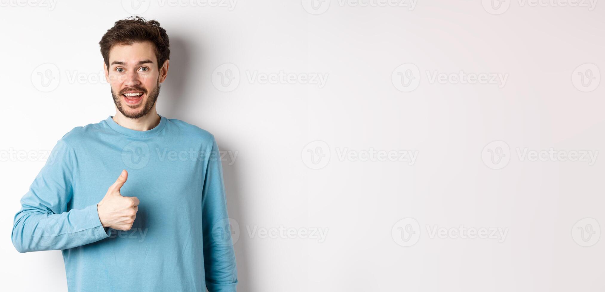 Cheerful young man agree and say yes, show thumb up in approval, smiling happy at camera, standing over white background photo