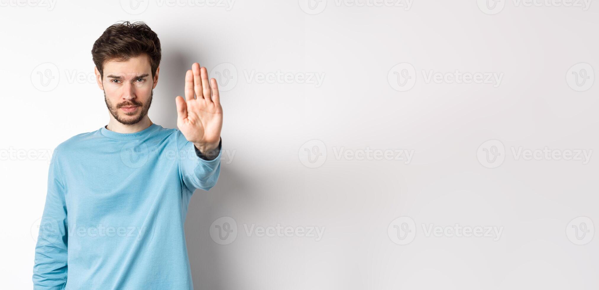 Serious young man looking confident with hand stretched out, saying stop, prohibit something bad or give warning, standing over white background photo