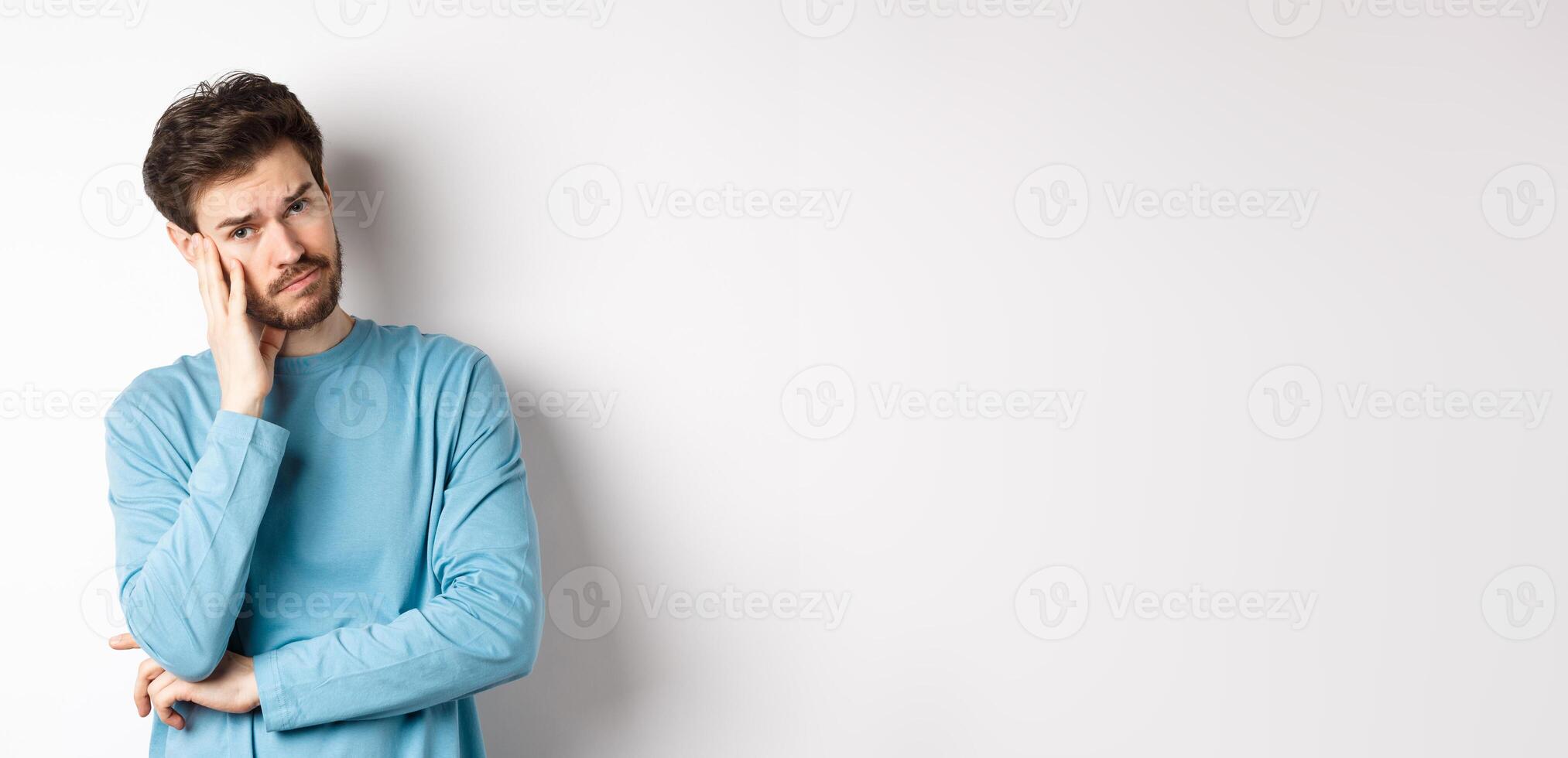 Sad young man looking frustrated, touching temple of head and frowning uneasy, standing against white background photo