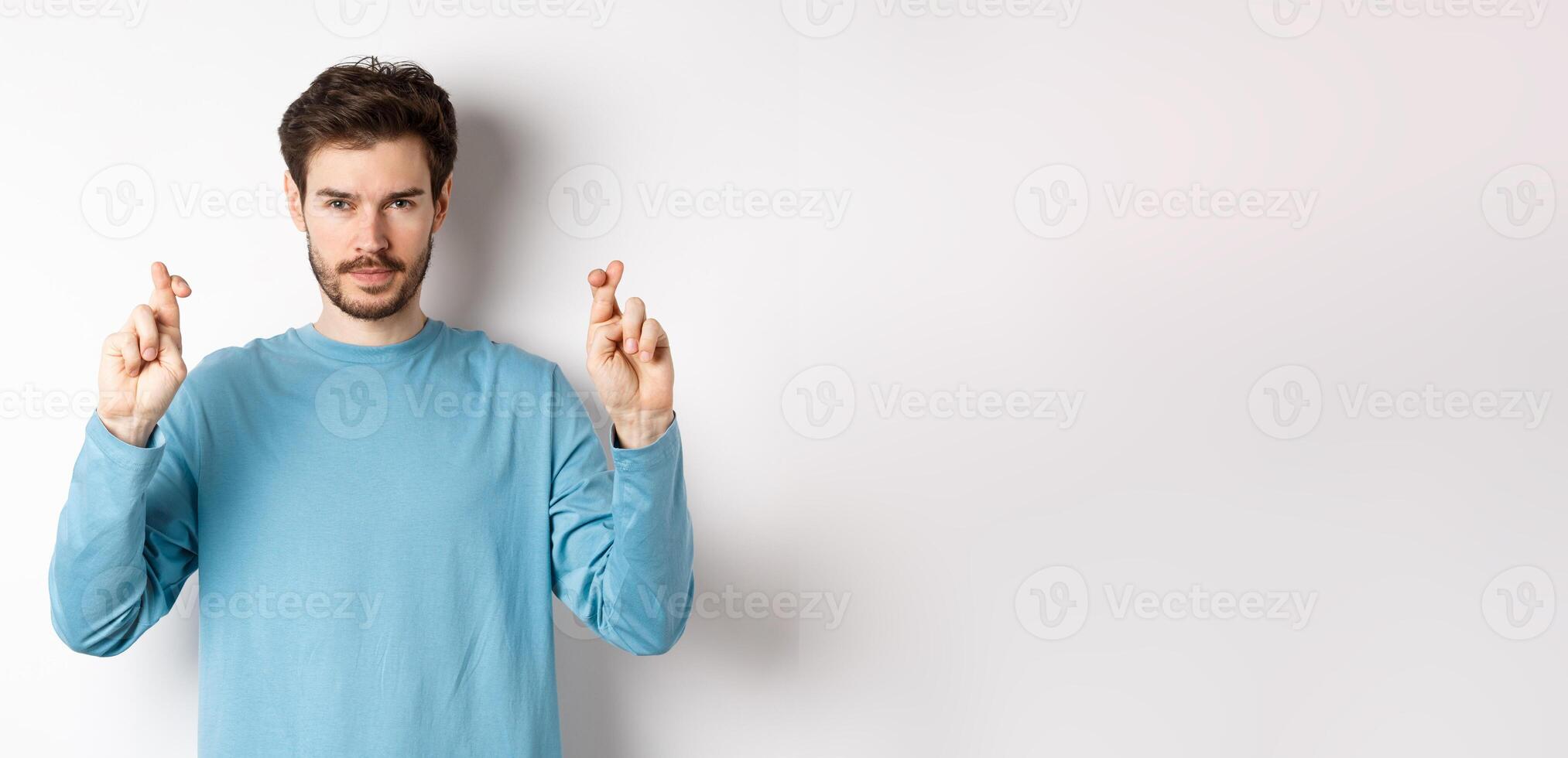 Hopeful and confident young man cross fingers for good luck, making wish or praying, anticipating results, standing over white background photo