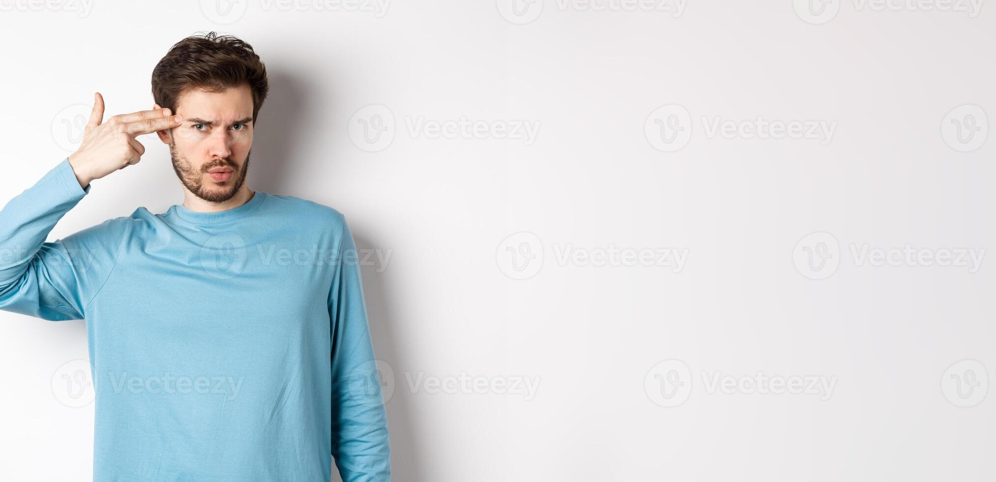 Annoyed young man frowning, showing shooting gun near head gesture, blow brain out sign, standing over white background photo