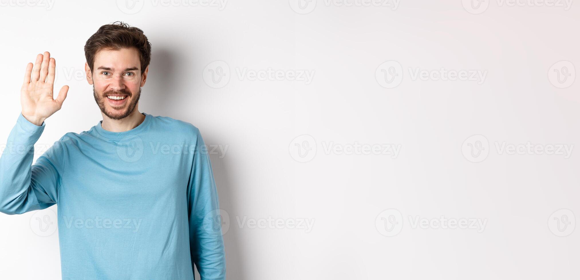 Cheerful young man with beard saying hello, looking friendly and waving hand to greet you, standing over white background photo